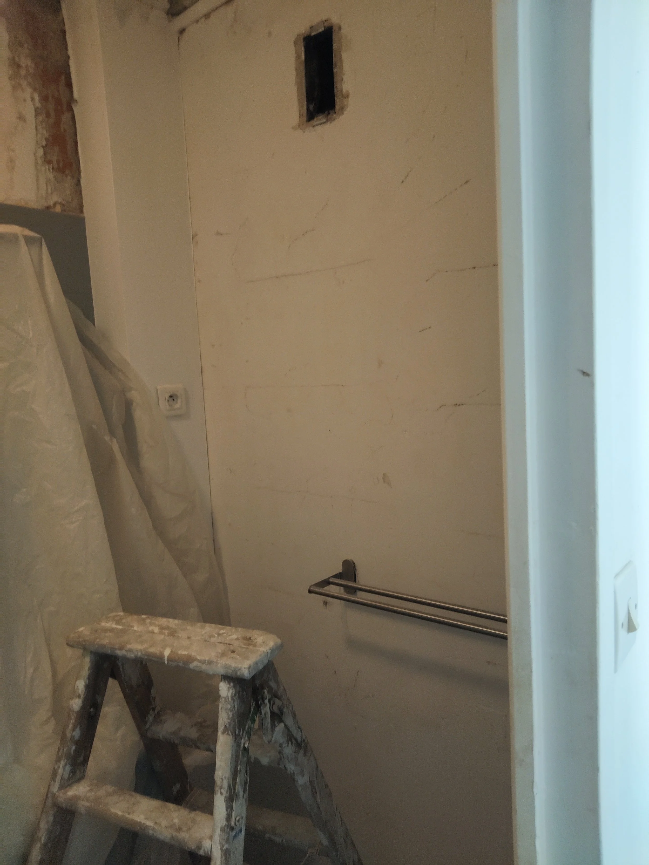 Une pièce en rénovation avec un échafaudage en bois, un mur en cours de peinture, une prise électrique, un porte-serviettes métallique, et une toile de projection ou un film plastique sur le sol.