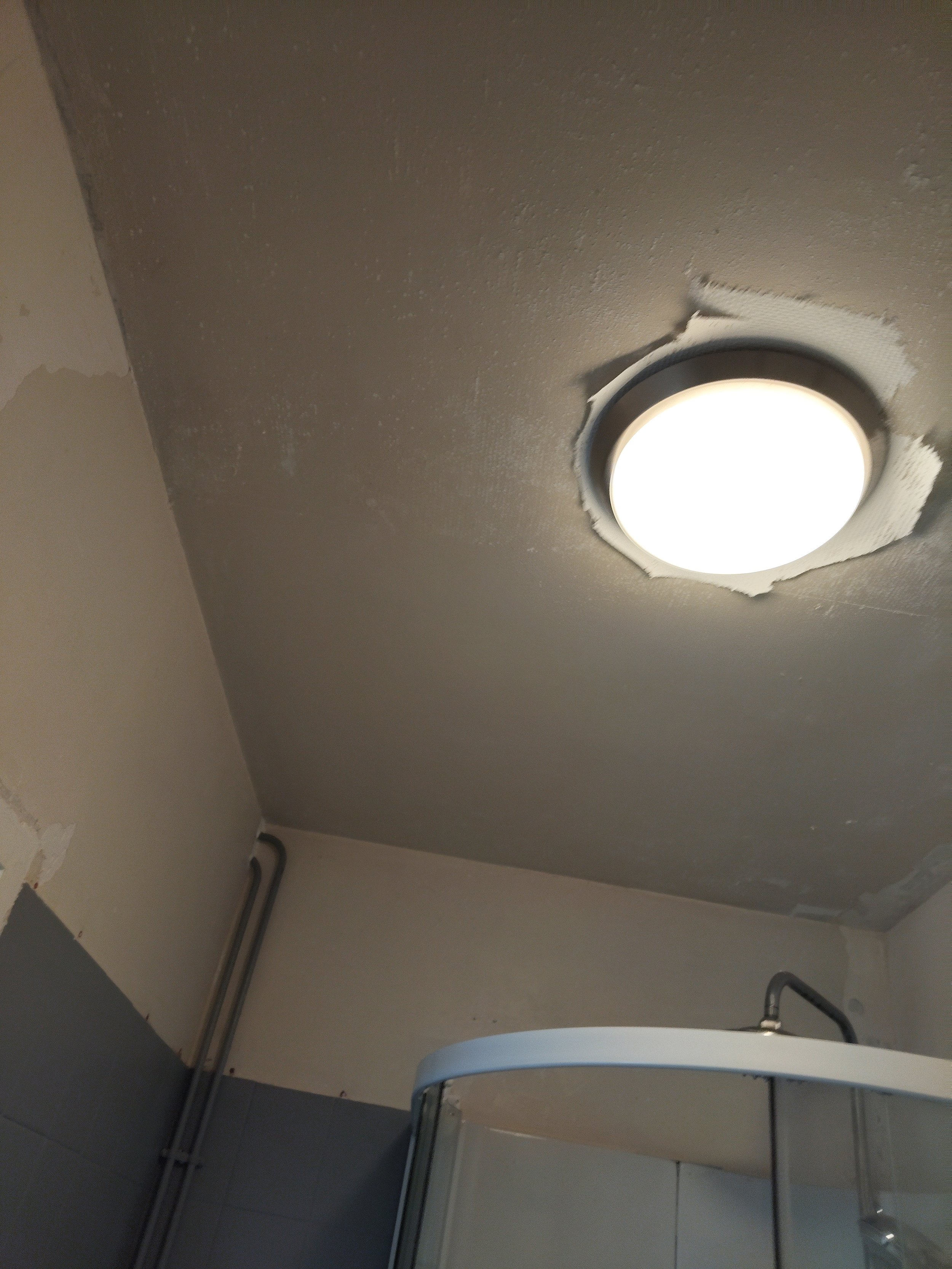Il y a un plafond avec un trou autour d'une lumière encastrée, des murs et des conduits électriques visibles en bas à gauche, et une chaise en zinc dans la partie inférieure de l'image.