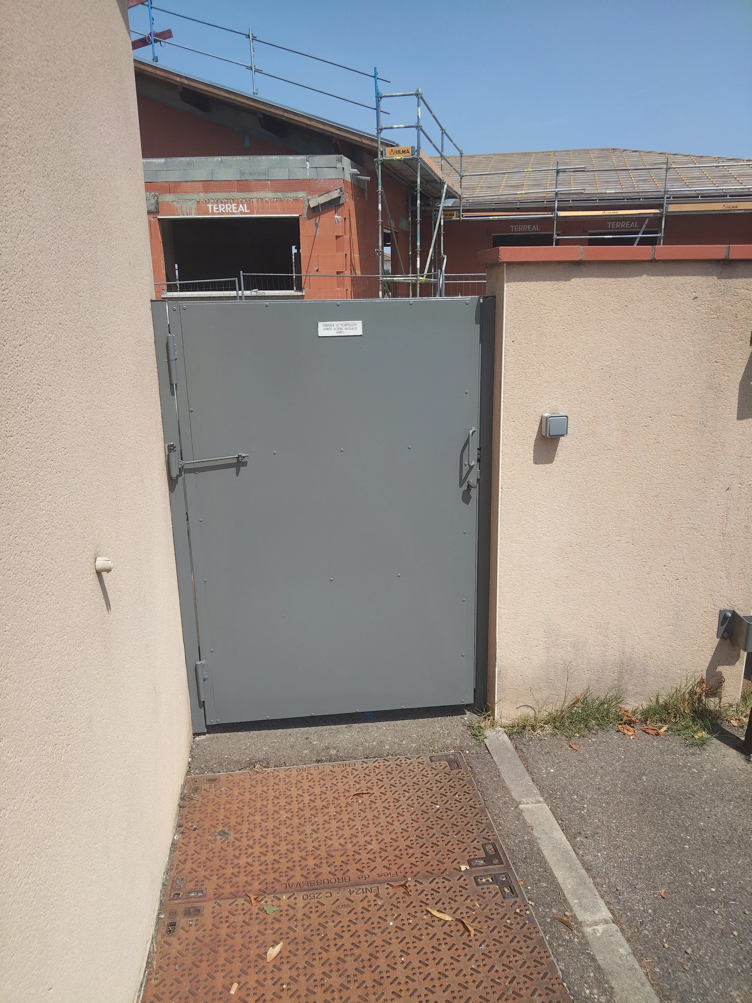 Une porte grise en métal avec un autocollant blanc, entourée de murs beige et une zone pavée avec une grille en fer et des feuilles mortes, devant un bâtiment en construction.