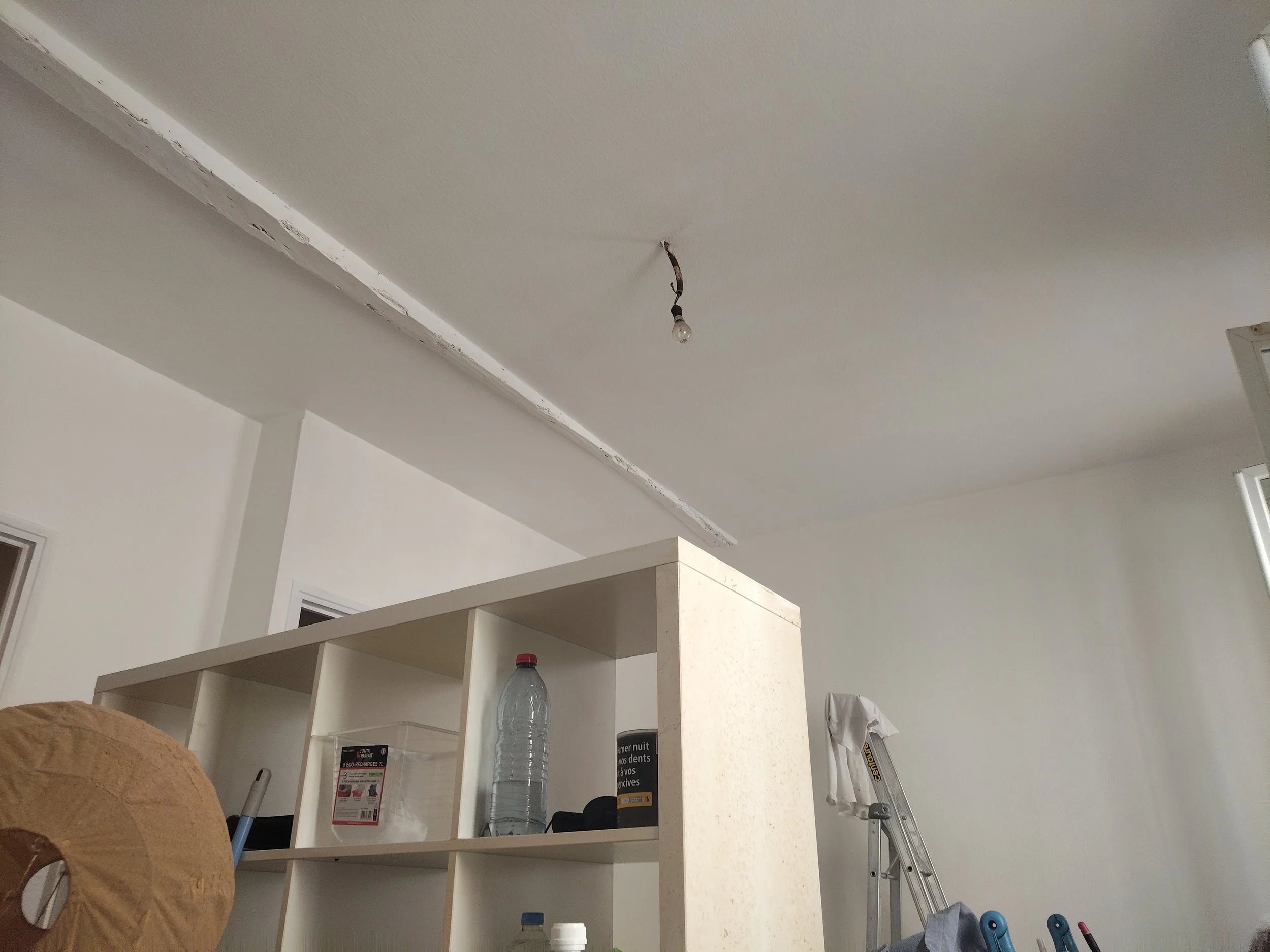 Plafond en train d'être réparé dans une pièce avec une lumière suspendue, étagères blanches avec divers objets, et une bouteille d'eau sur un plan de travail en marbre.