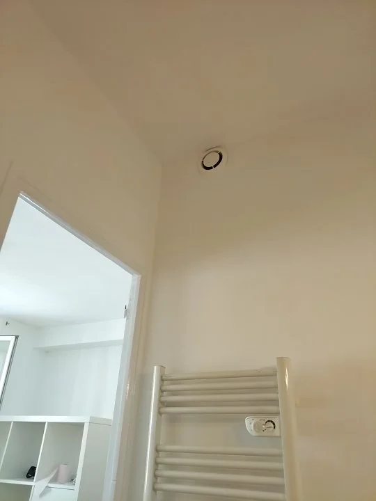 Intérieur d'une pièce avec un plafond, un mur blanc, un radiateur blanc mural, une prise électrique, et une porte donnant sur un autre espace lumineux.