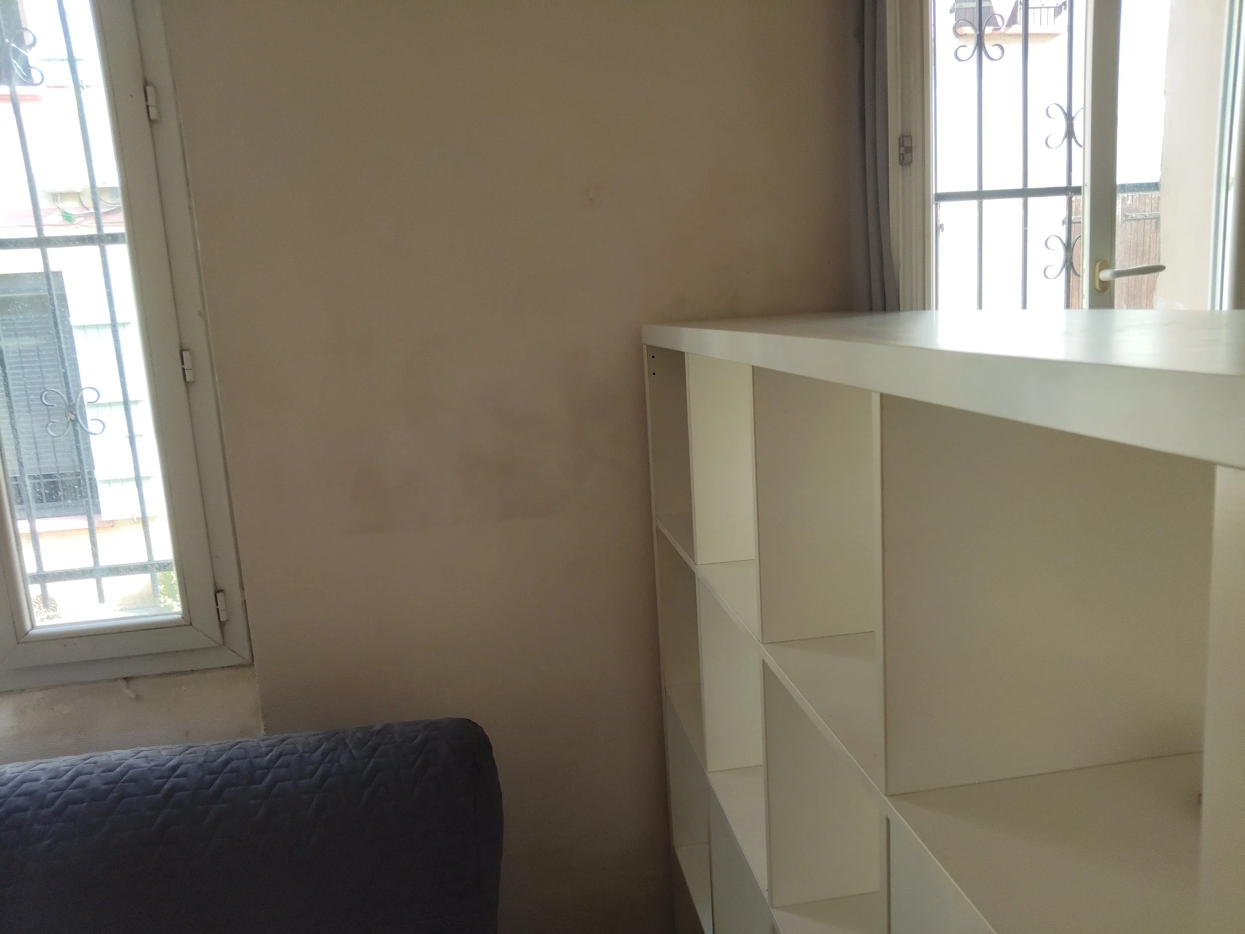 Une pièce avec une fenêtre, un mur beige, une étagère blanche vide et un lit sombre au premier plan.