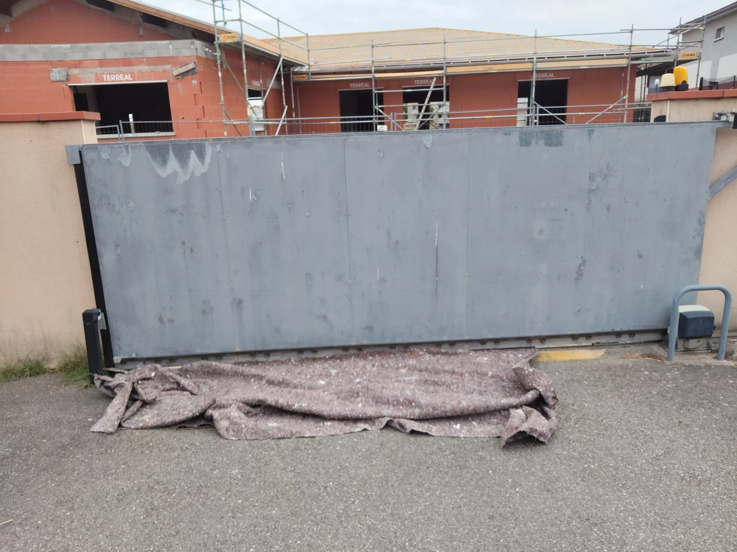 Une clôture métallique en face d'une maison en construction avec des échafaudages autour, une grande porte en métal en premier plan et des débris au sol.