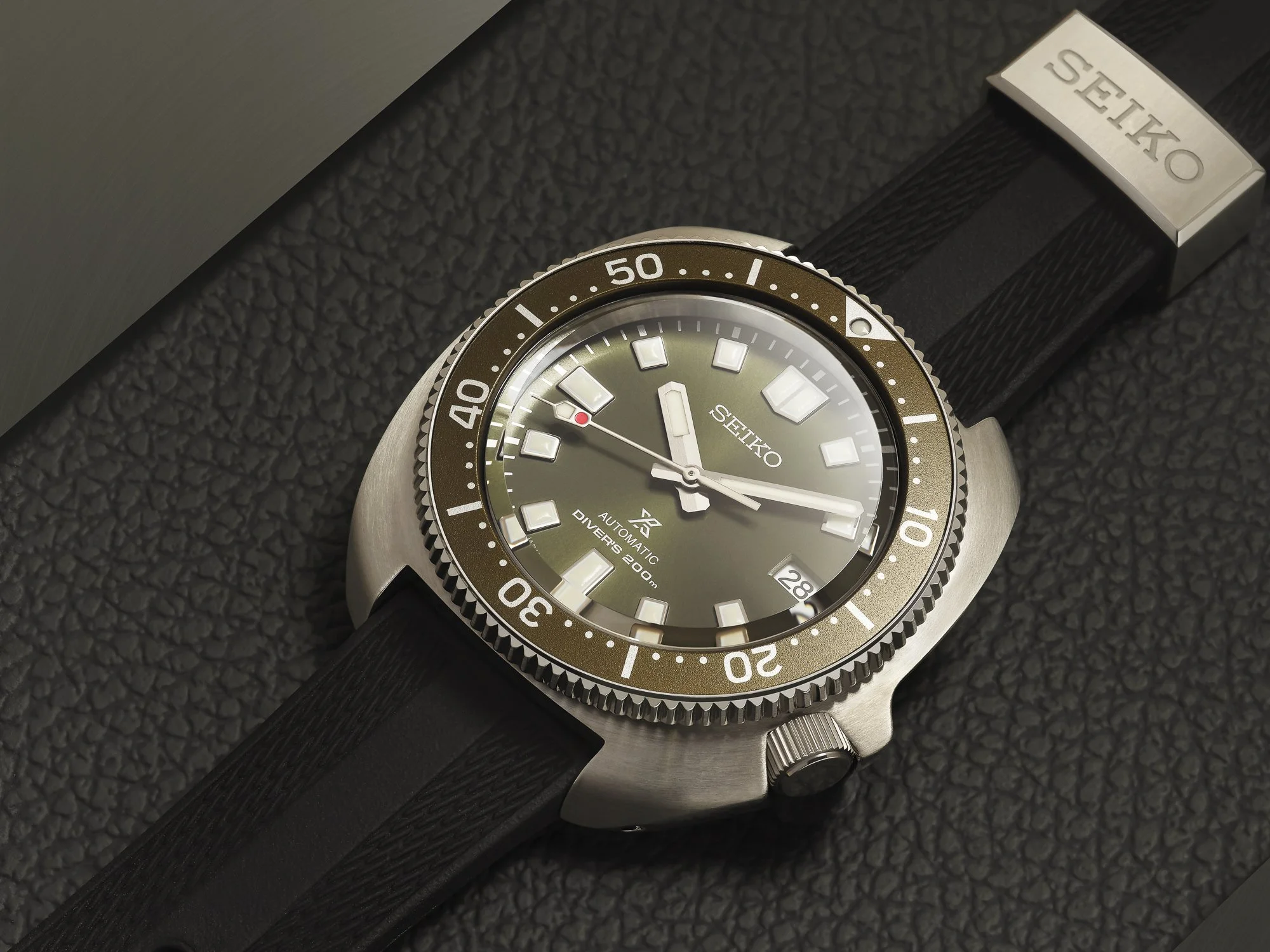 Een Seiko duikhorloge met een zwarte band en een zilveren kast, liggend op een zwarte, ruw oppervlak.