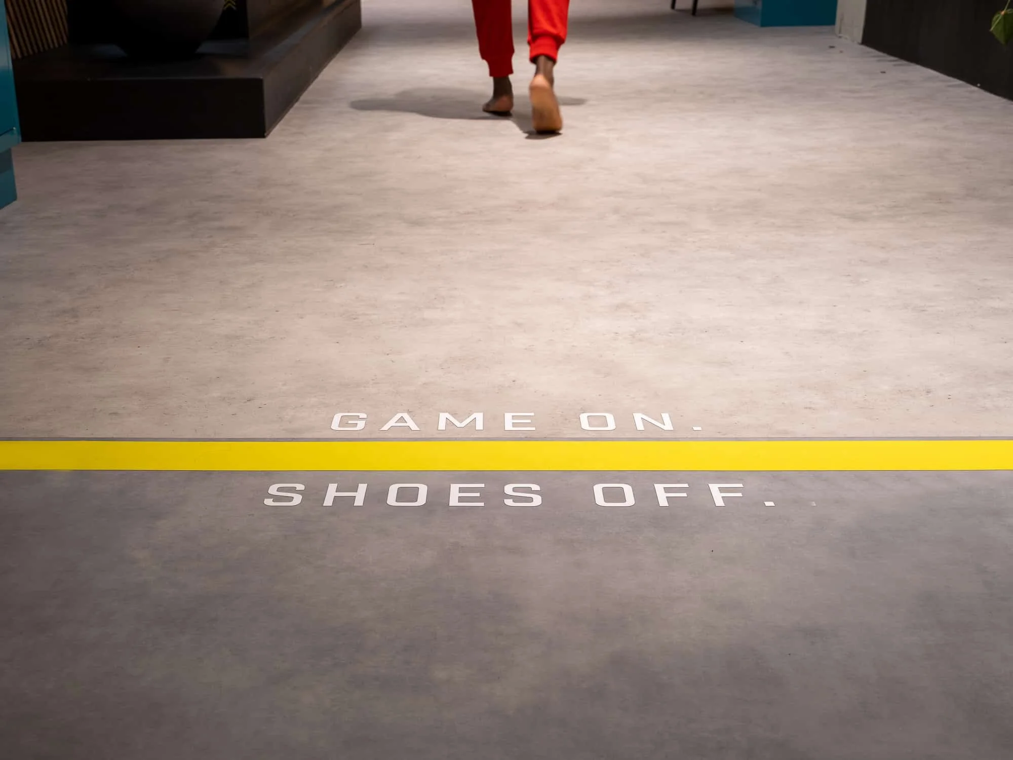 Een vloer met tekst die zegt 'GAMES ON, SHOES OFF' en een persoon die op blote voeten in de achtergrond loopt.