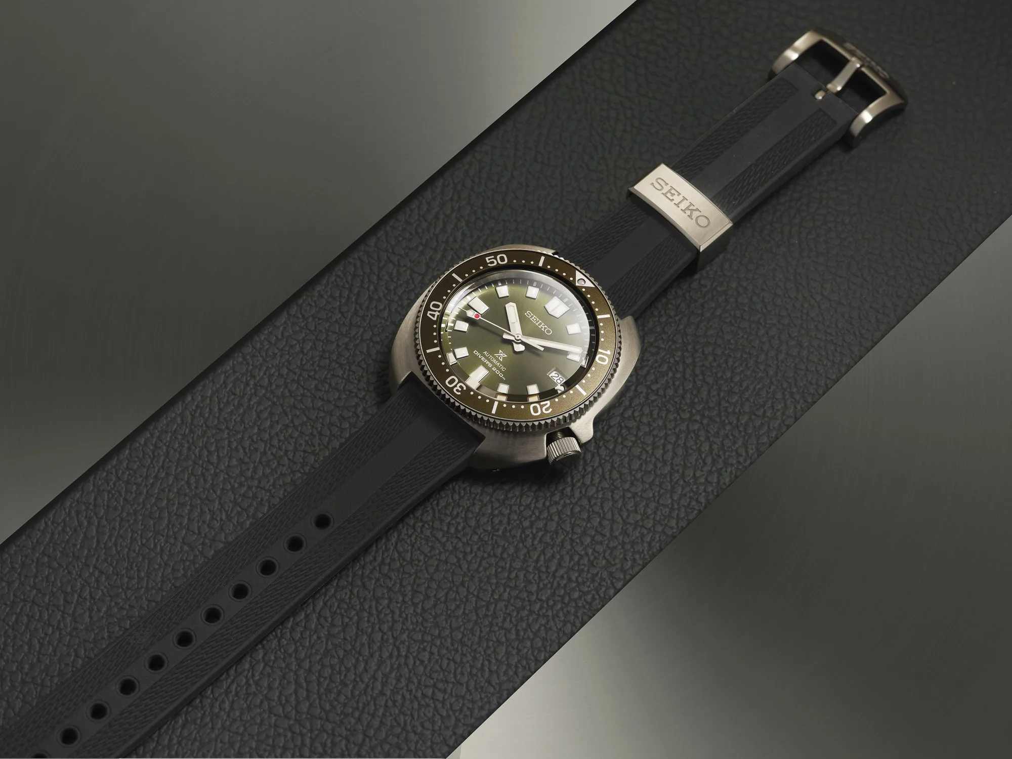 Een Seiko duikhorloge met een zwarte band en een zwarte wijzerplaat ligt op een zwarte, textured ondergrond.