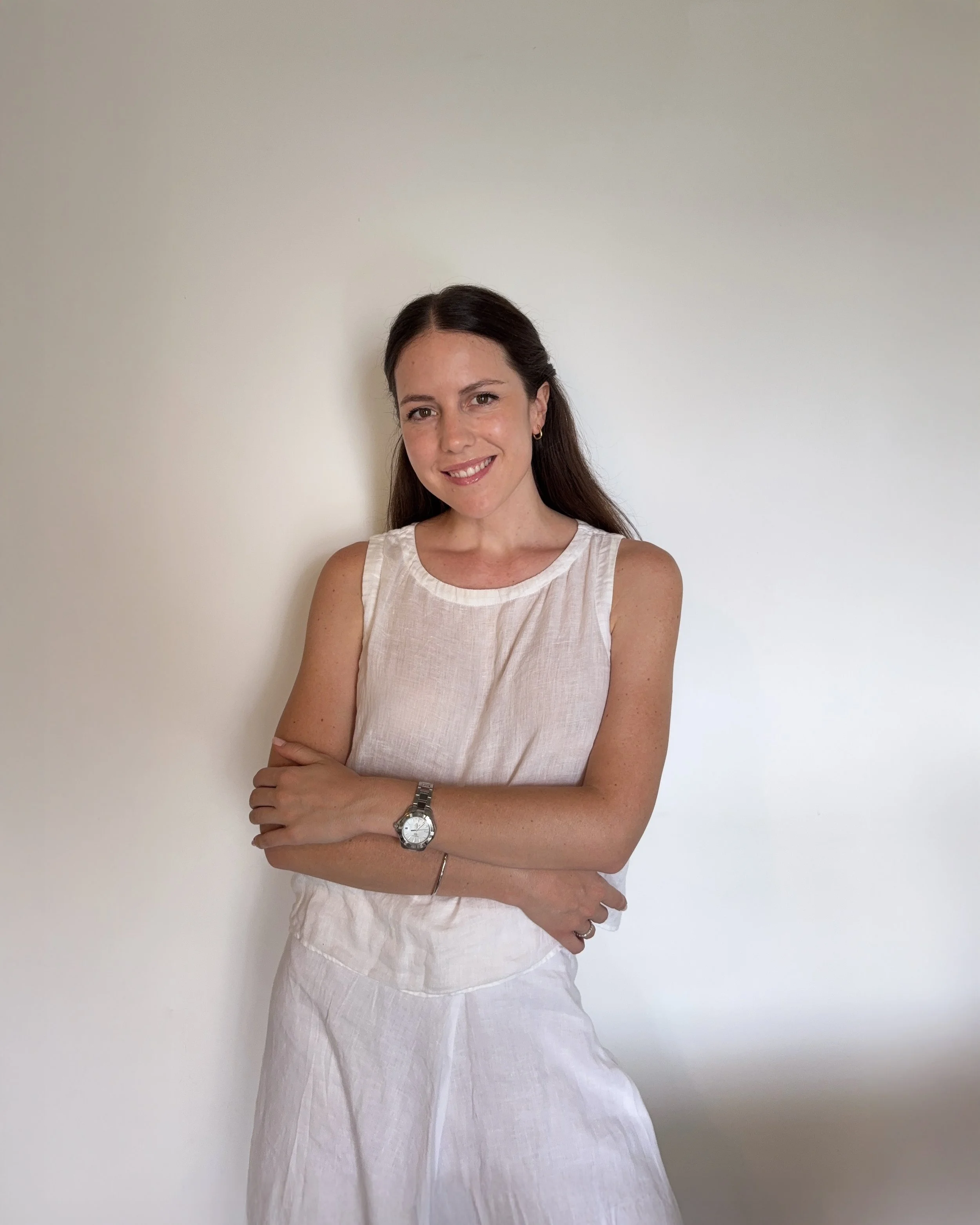 Una mujer sonriente con piel clara, cabello castaño oscuro y pantalones blancos de lino, lleva un reloj plateado en la muñeca izquierda y un arete pequeño en la oreja derecha, posando con los brazos cruzados frente a una pared blanca.