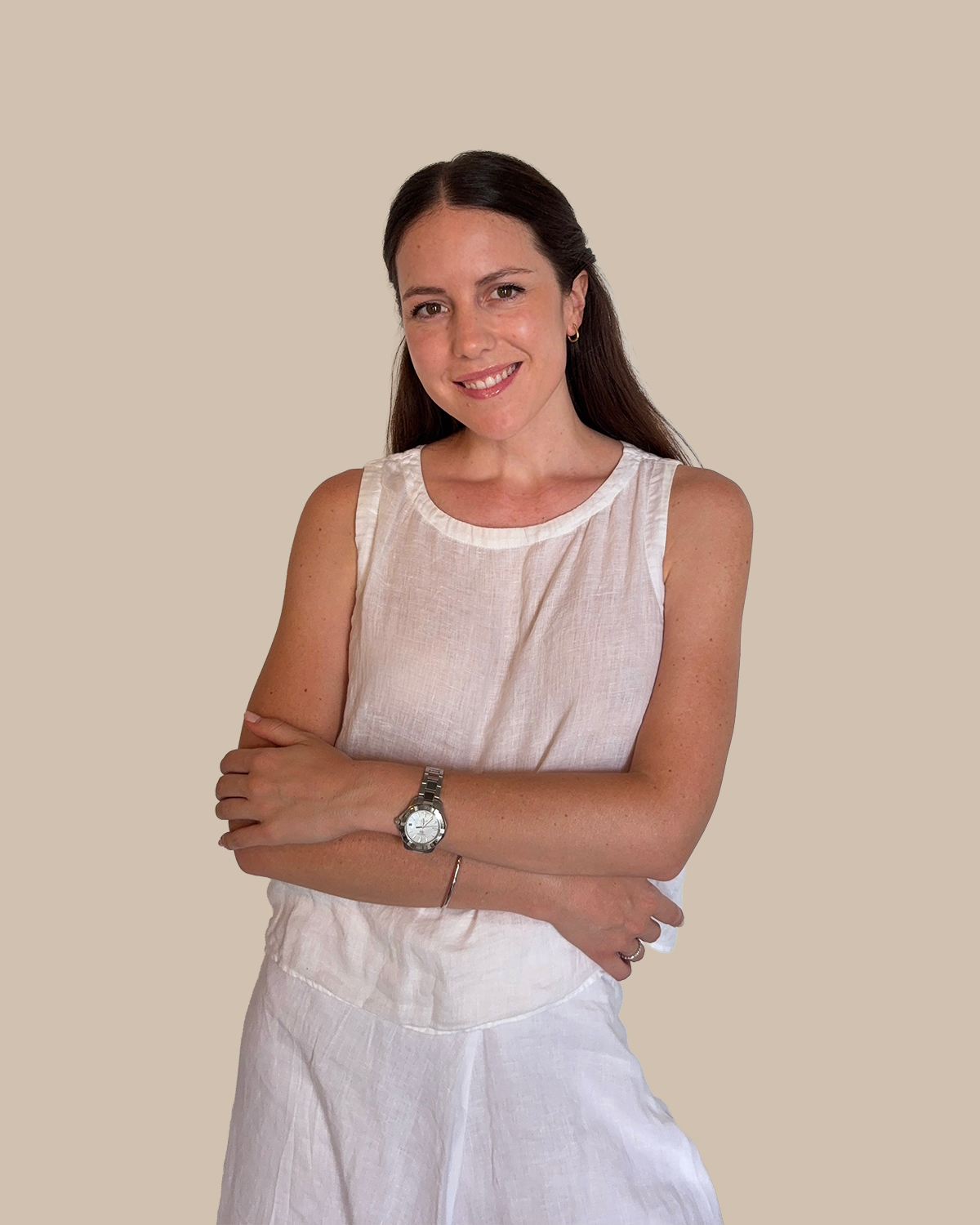 A woman with long dark hair smiling, wearing a sleeveless white top and white skirt, with her arms crossed, standing against a beige background.