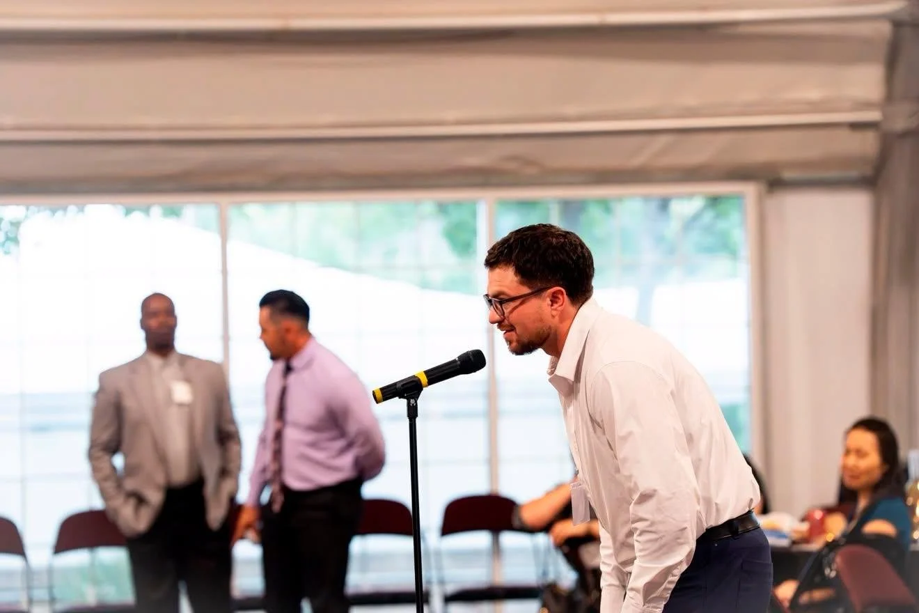 A man in a white shirt and glasses bowing at a microphone during a conference or event inside a large tent with several attendees in the background.