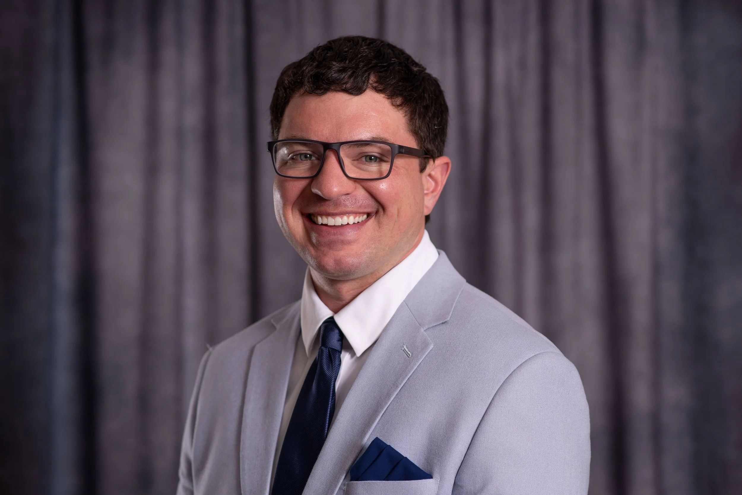 A man wearing glasses, a light gray suit, white shirt, and dark tie smiling against a gray curtain background.