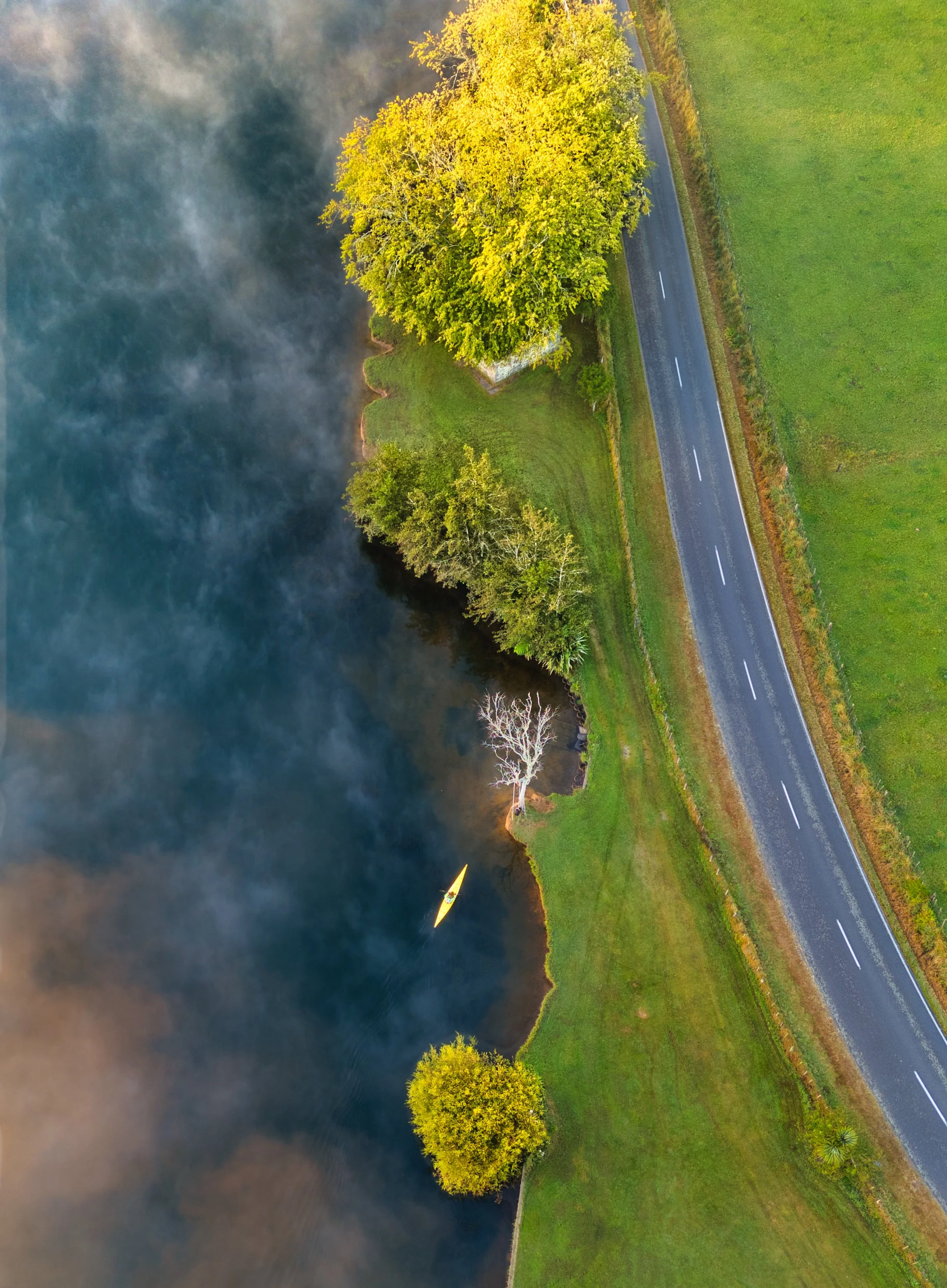 A small yellow boat is on a dark, smoke-filled river, with tall green trees along the riverbank and a road with green grass and trees running parallel to the river.