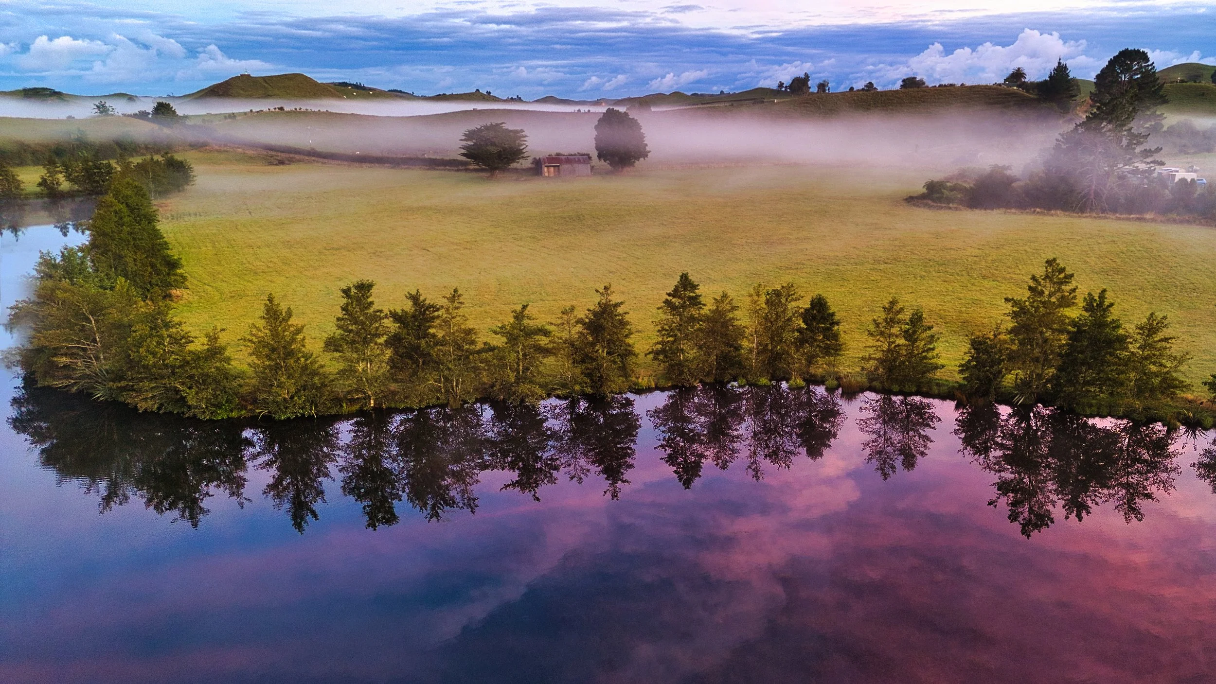 Scenic landscape with a calm river reflecting trees and a distant grassy hill, fog drifting through the area, with a partly cloudy sky overhead.