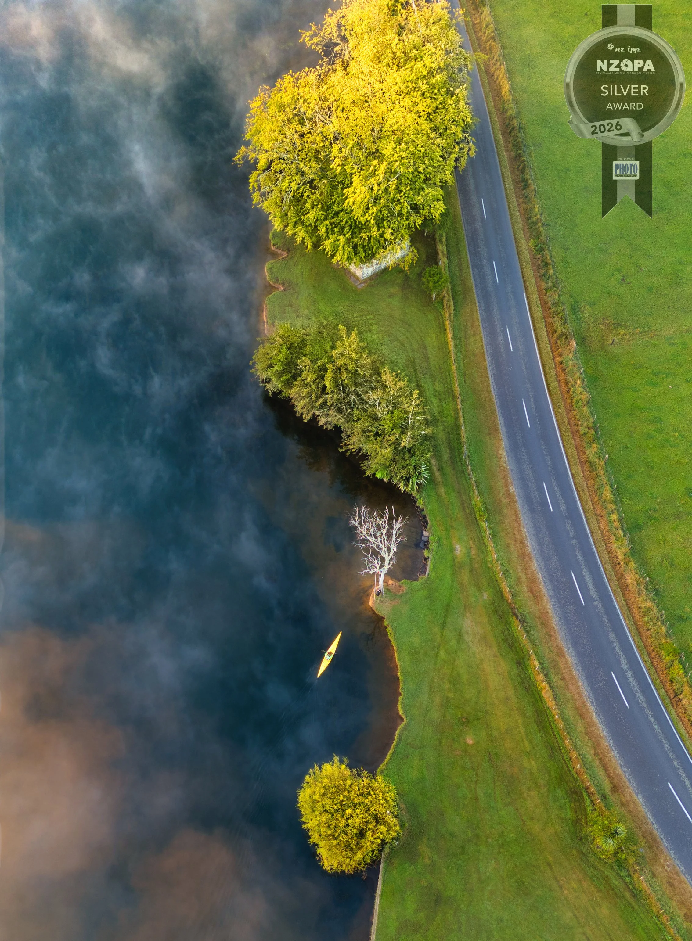 Aerial view of a river with dark water and smoke rising from the bank, trees along the shoreline, and a road running parallel to the river.
