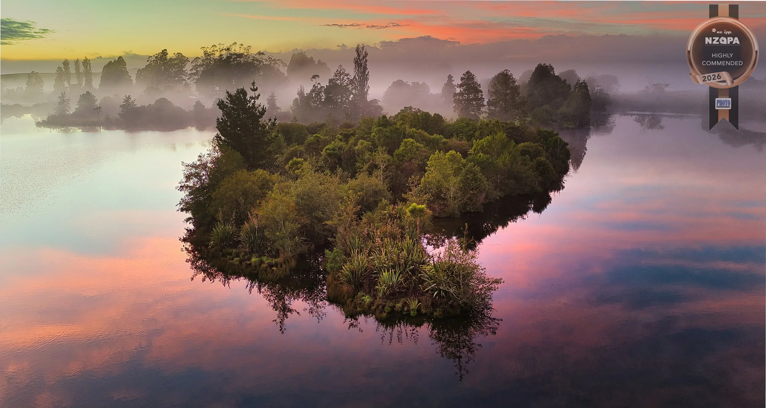 A serene landscape featuring a lake with reflections of trees and a colorful, misty sky at sunrise or sunset. There is a small, densely wooded island in the middle of the lake and a badge in the upper right corner indicating it was highly commended i