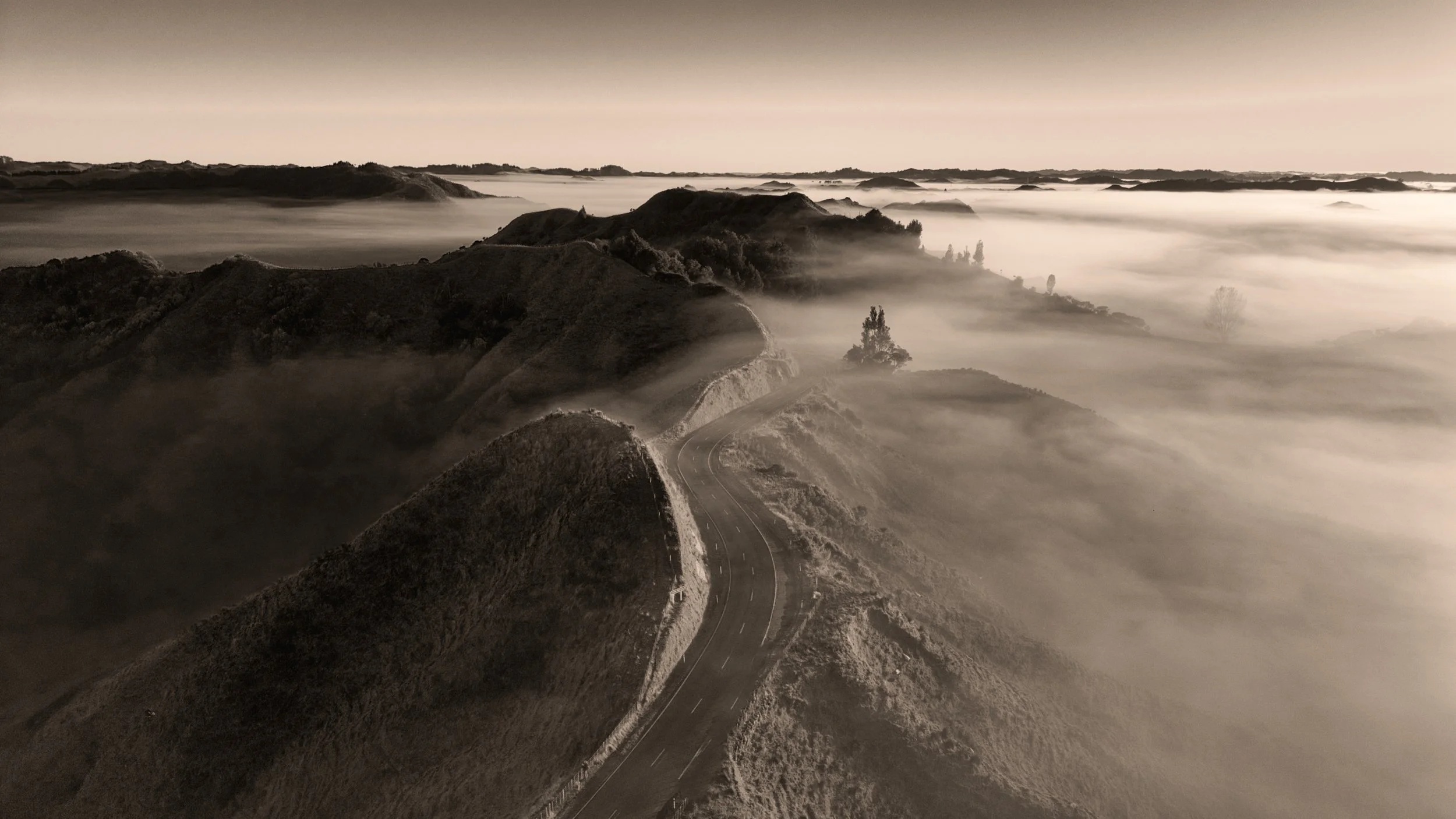 A grayscale landscape of rolling hills and mountains with a winding road, surrounded by fog or mist.