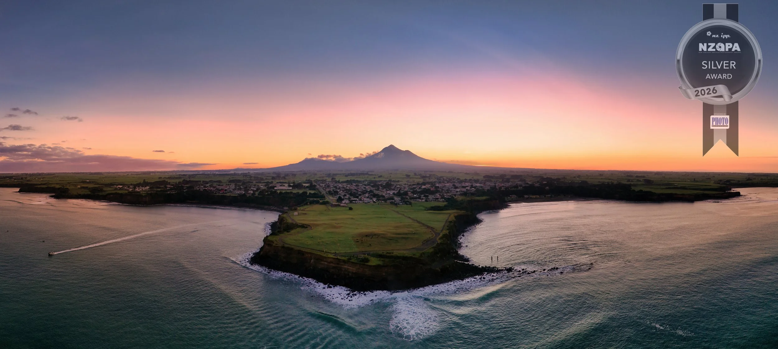 A scenic aerial view of a body of water with a small peninsula, green fields, and a town at sunset, with a mountain in the background.