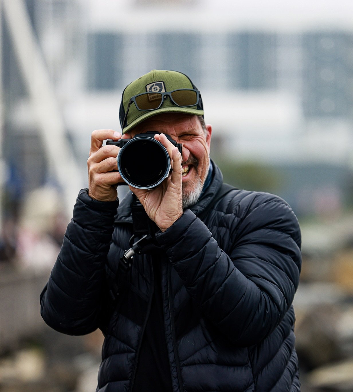 Emile Grundlingh taking a photo with a camera outdoors, wearing a black jacket and a green cap with sunglasses on top.