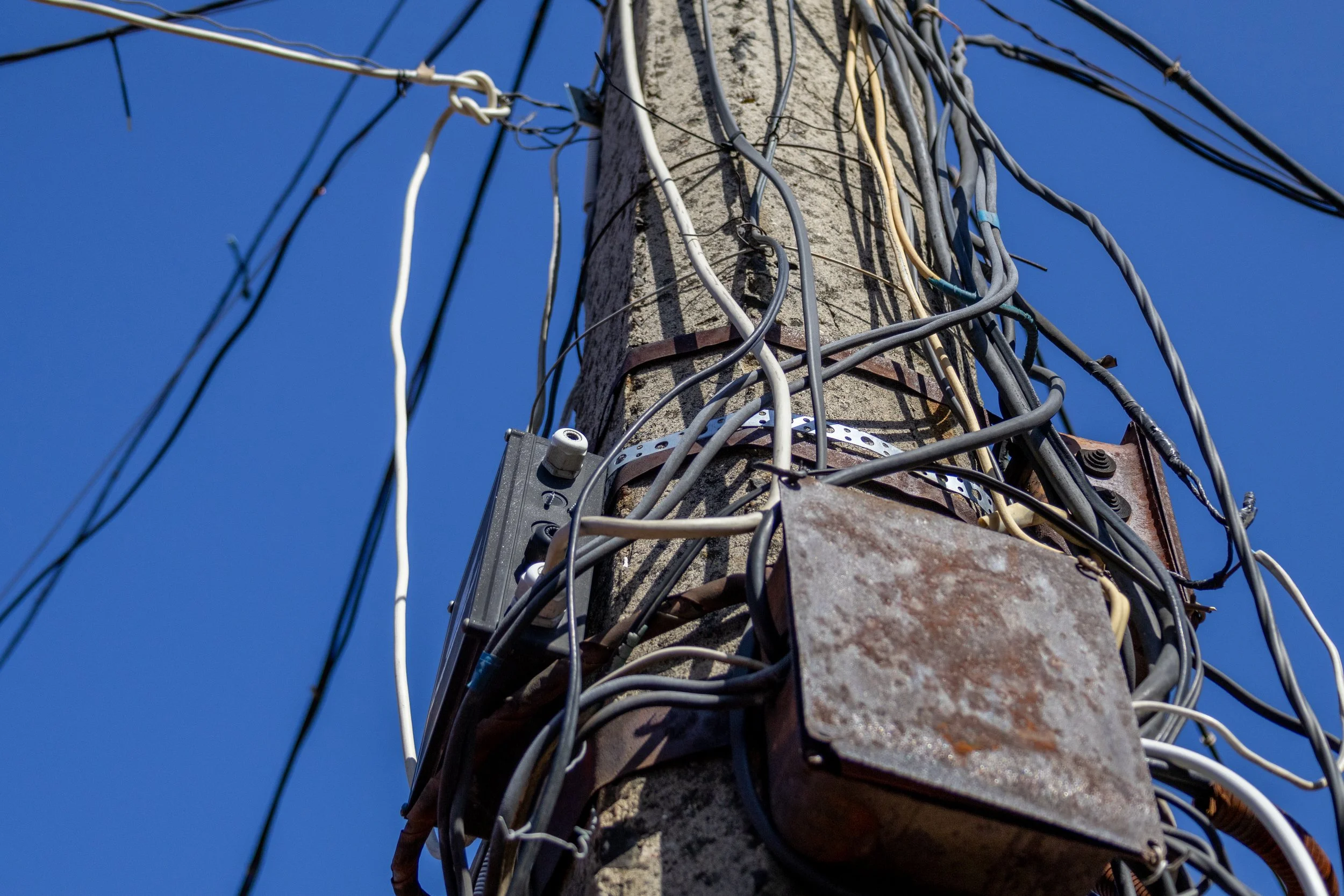 Close-up of a utility pole with numerous tangled wires and cables against a clear blue sky.