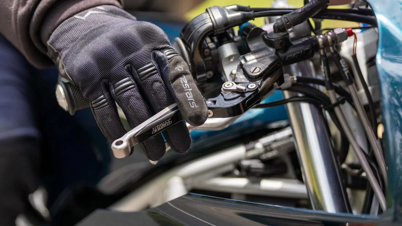 Close-up of a motorcyclist's hand wearing a black glove, gripping the handlebar of a motorcycle, with a visible wrench near the hand.