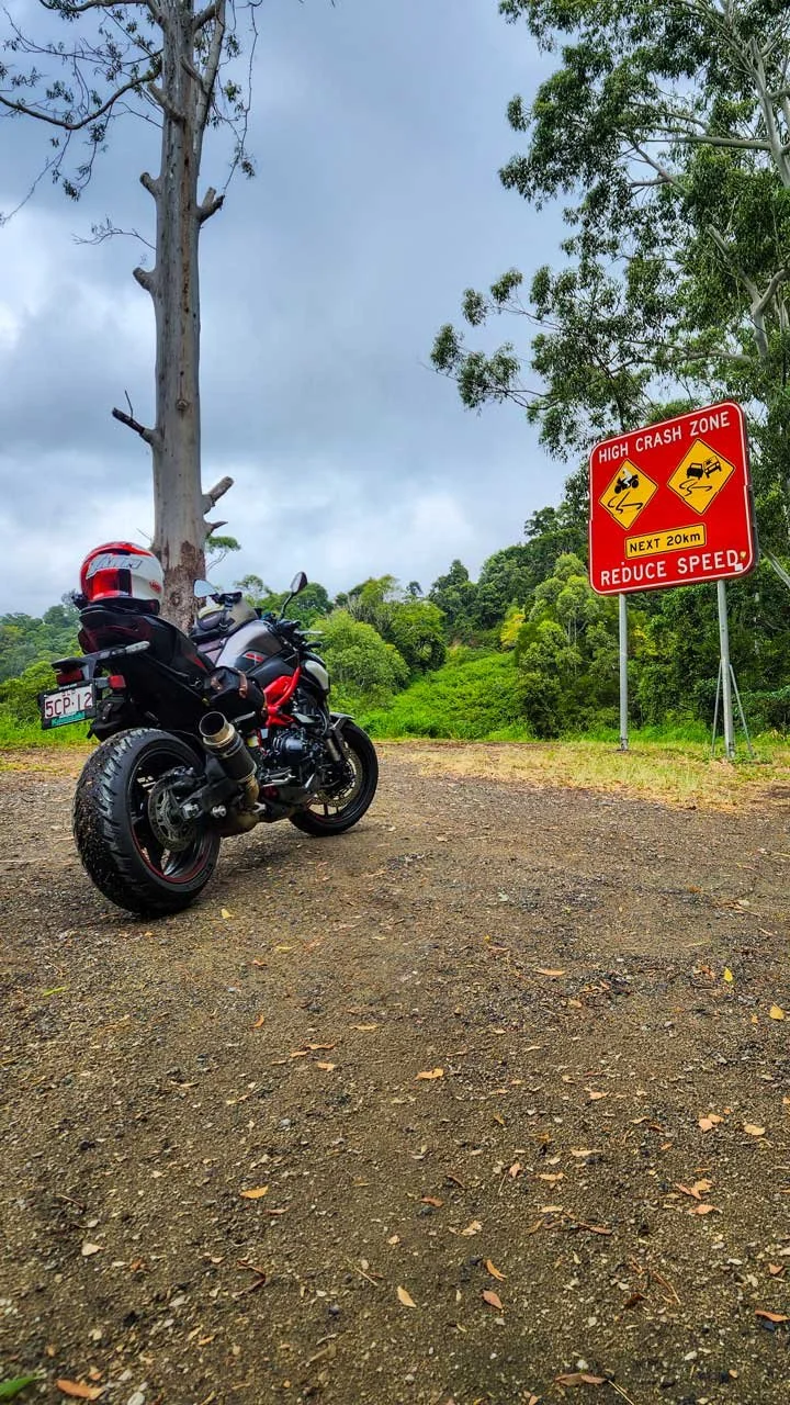 A motorcycle parked on a dirt patch next to a bare tree and a lush green forest, with a red warning sign indicating a high crash zone ahead and urging drivers to reduce speed.