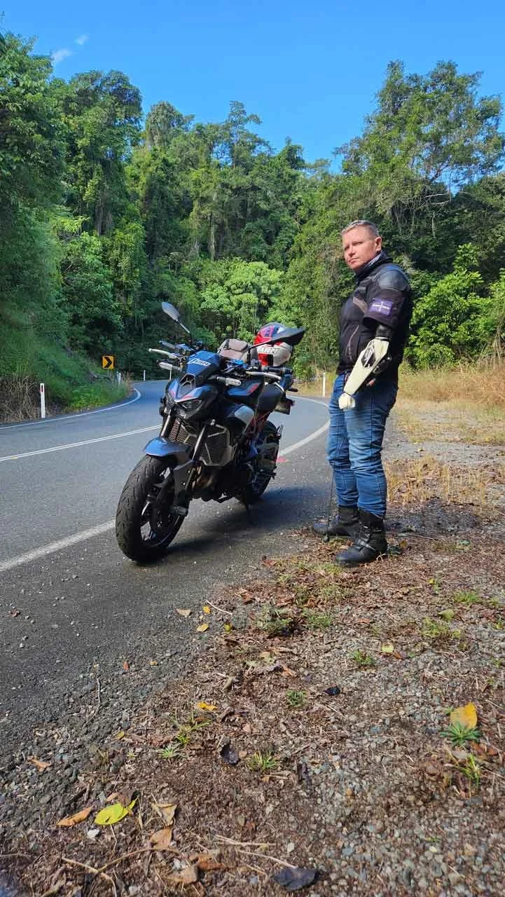 A man wearing motorcycle gear standing next to a black motorcycle on a winding road surrounded by green trees.