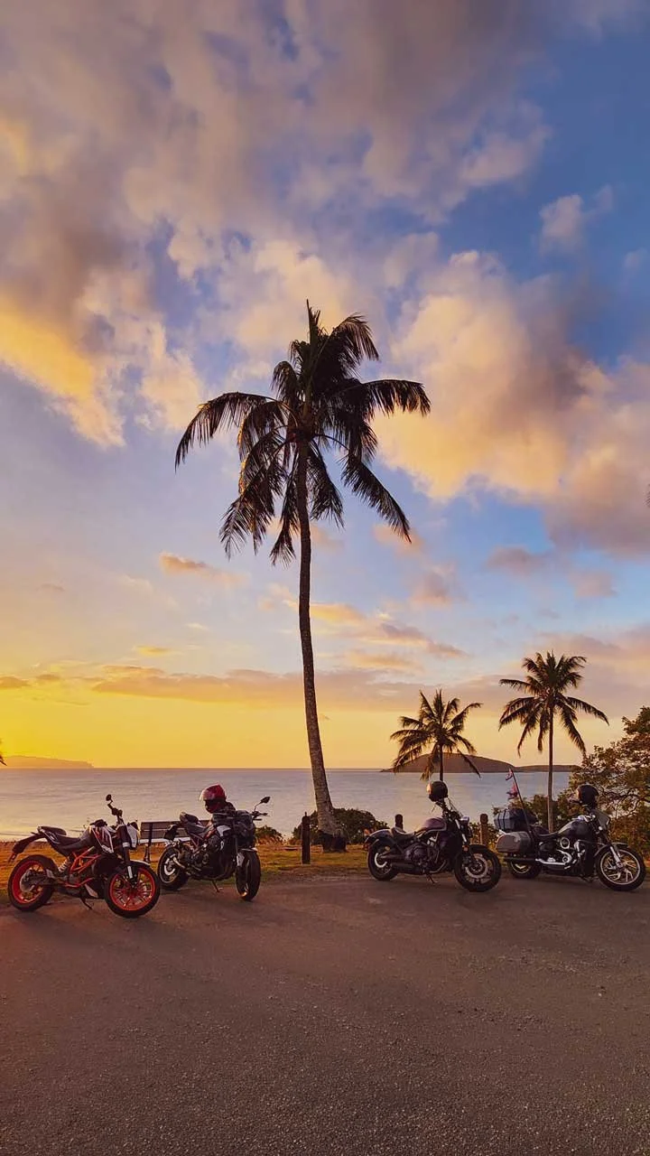 Four motorcycles parked along a road near a beach with palm trees and a sunset sky with clouds.
