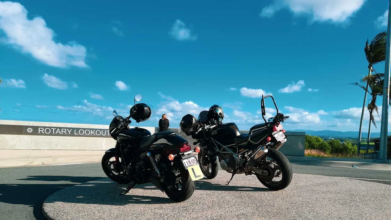 Two black motorcycles with helmets parked on a scenic lookout with a sign that reads "Rotary Lookout" under a blue sky with clouds and palm trees in the background.