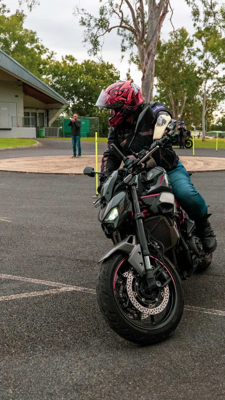 A motorcyclist wearing a pink helmet and black riding gear, sitting on a black and gray motorcycle in a parking lot with trees and a building in the background.