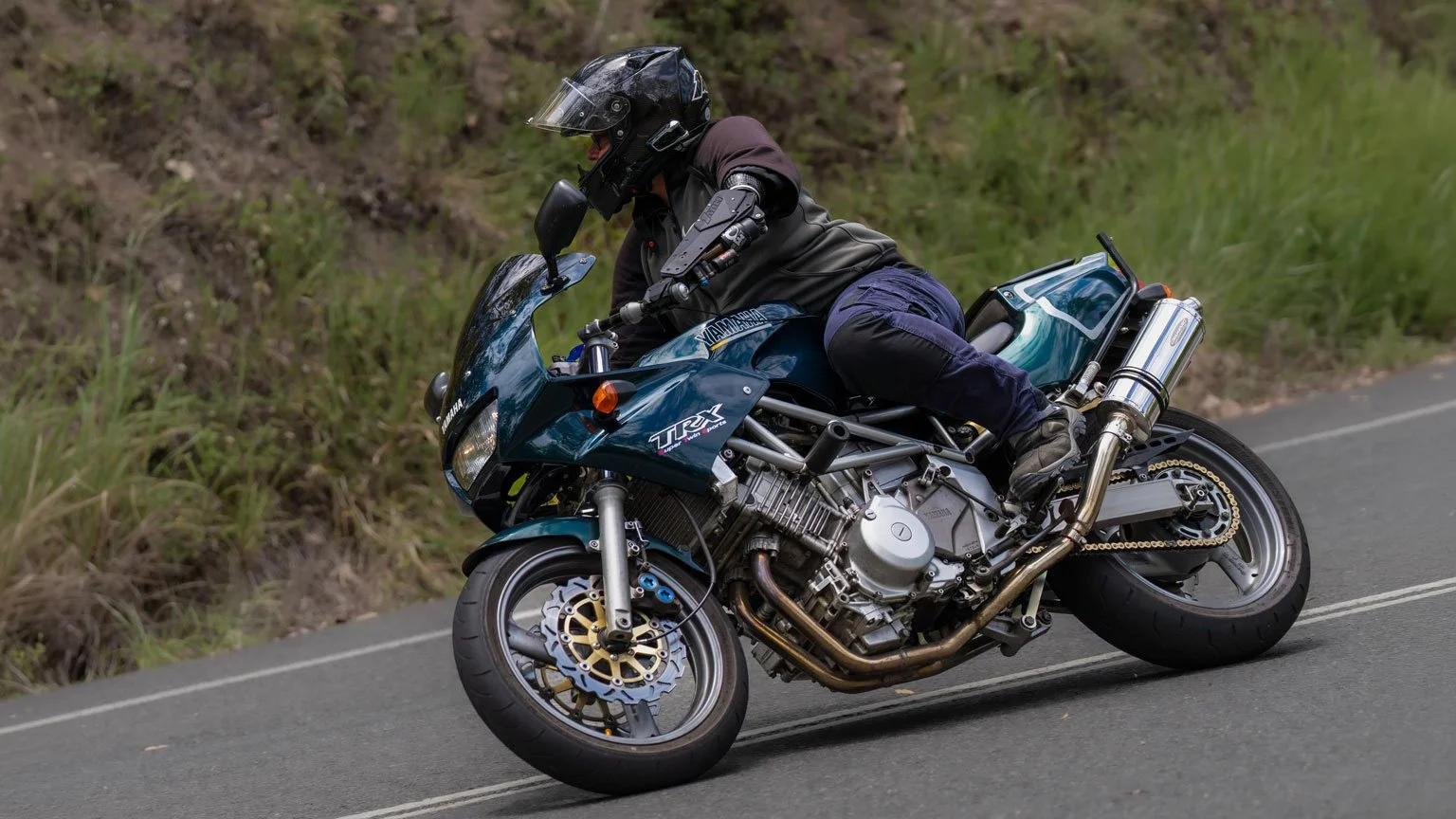 Motorcyclist wearing black helmet and gear leaning into a turn on a blue sport motorcycle on a paved countryside road with green foliage.