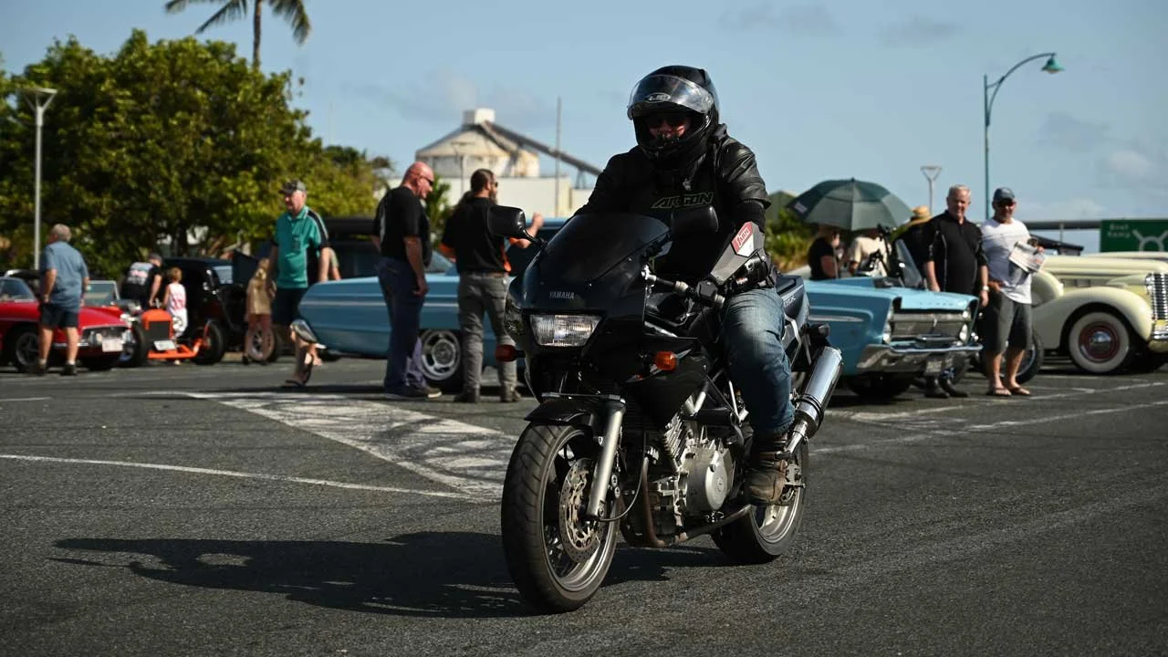 A man wearing a black helmet and leather jacket riding a black motorcycle in a parking lot with classic cars and people in the background.