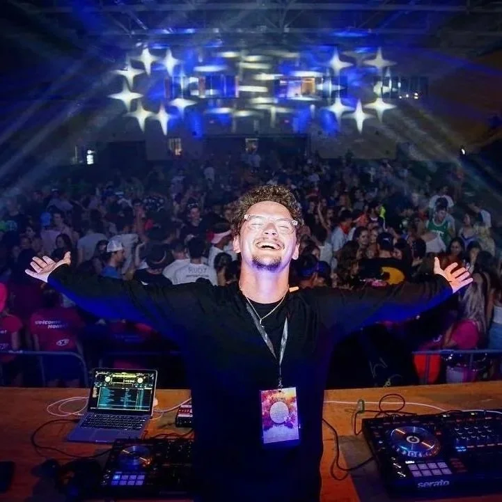 DJ smiling with arms outstretched at a crowded indoor music event with colorful lights.
