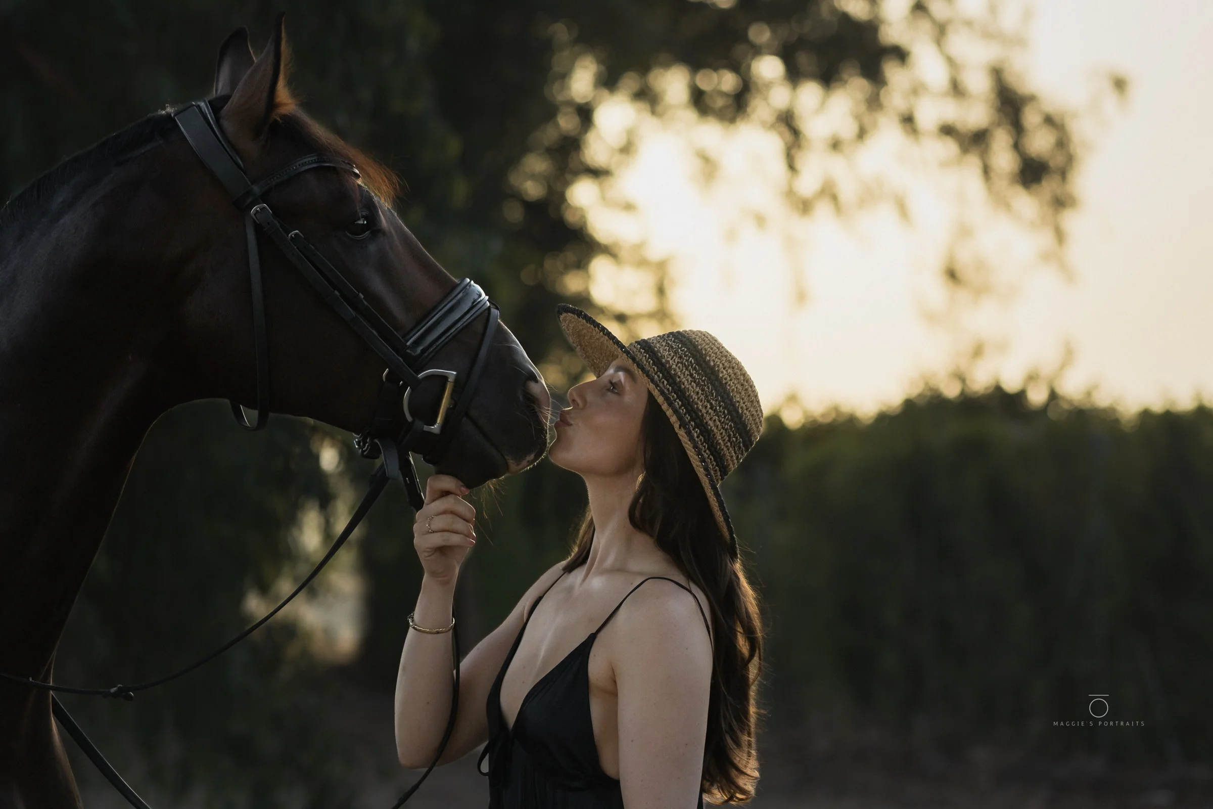 A woman with long, dark hair wearing a wide-brimmed straw hat and a black spaghetti strap dress gazes upward at a large dark brown horse with a glossy coat. The horse is wearing a bridle, and they are outdoors during sunset, with blurred trees and wa