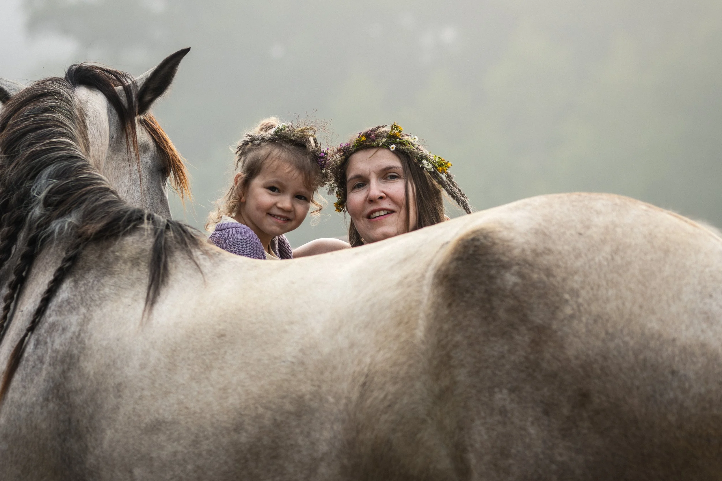 A woman and a young girl with flower crowns on their heads, smiling and leaning on a white horse, with a cloudy sky in the background.