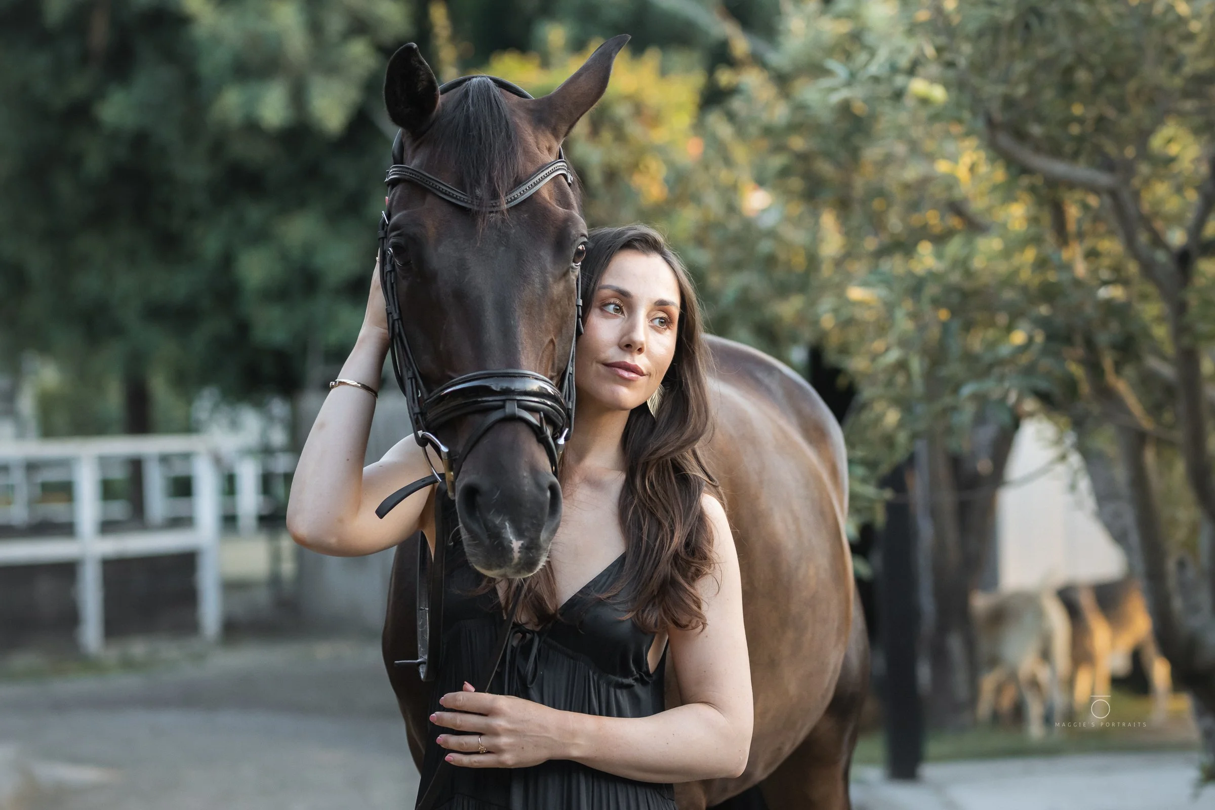 A woman with long brown hair wearing a black dress, standing next to a brown horse with a black mane, outdoors with trees in the background.