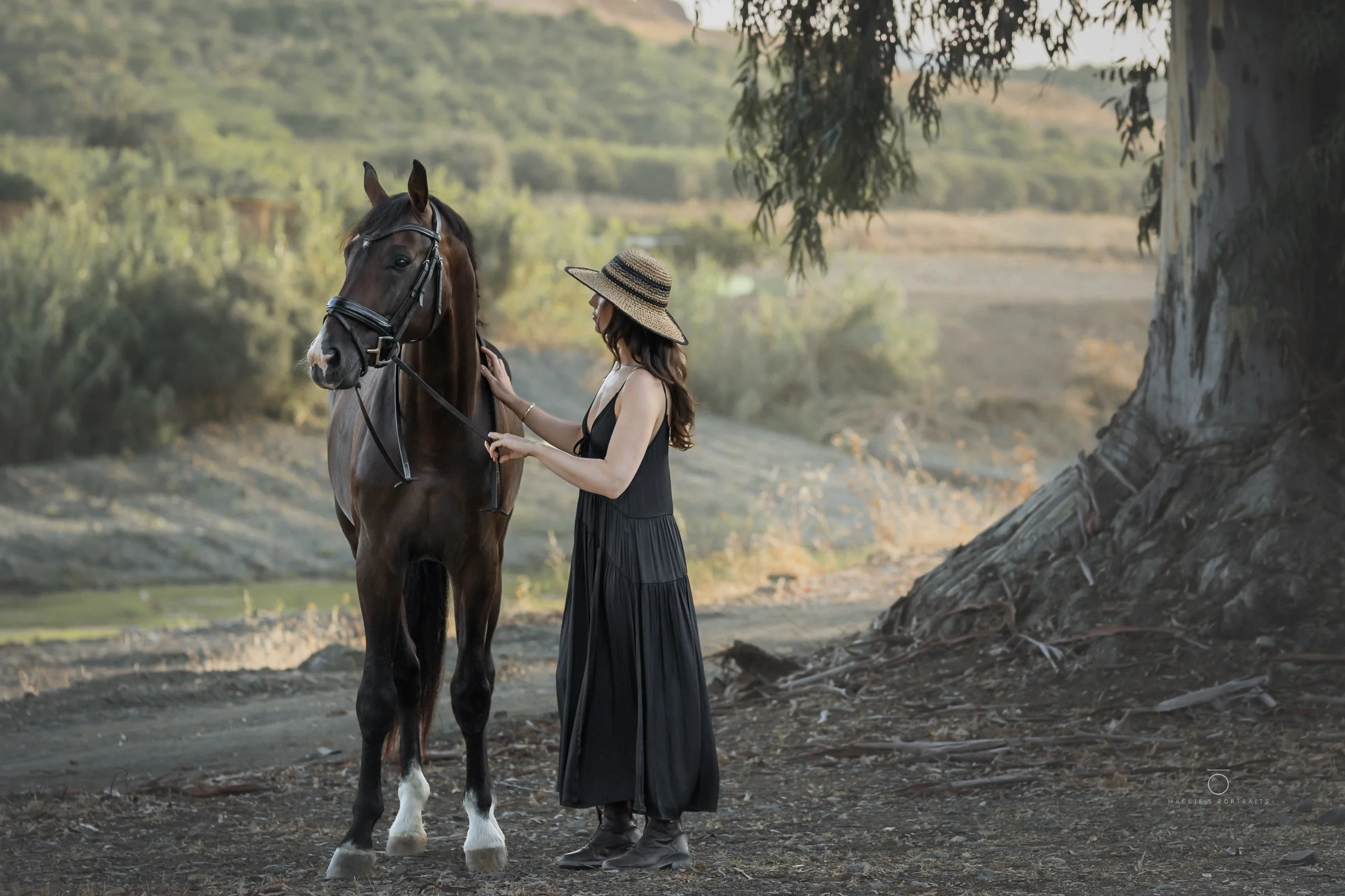 A woman wearing a straw hat and a black dress standing next to a brown horse near a large tree and a body of water in a rural outdoor setting.
