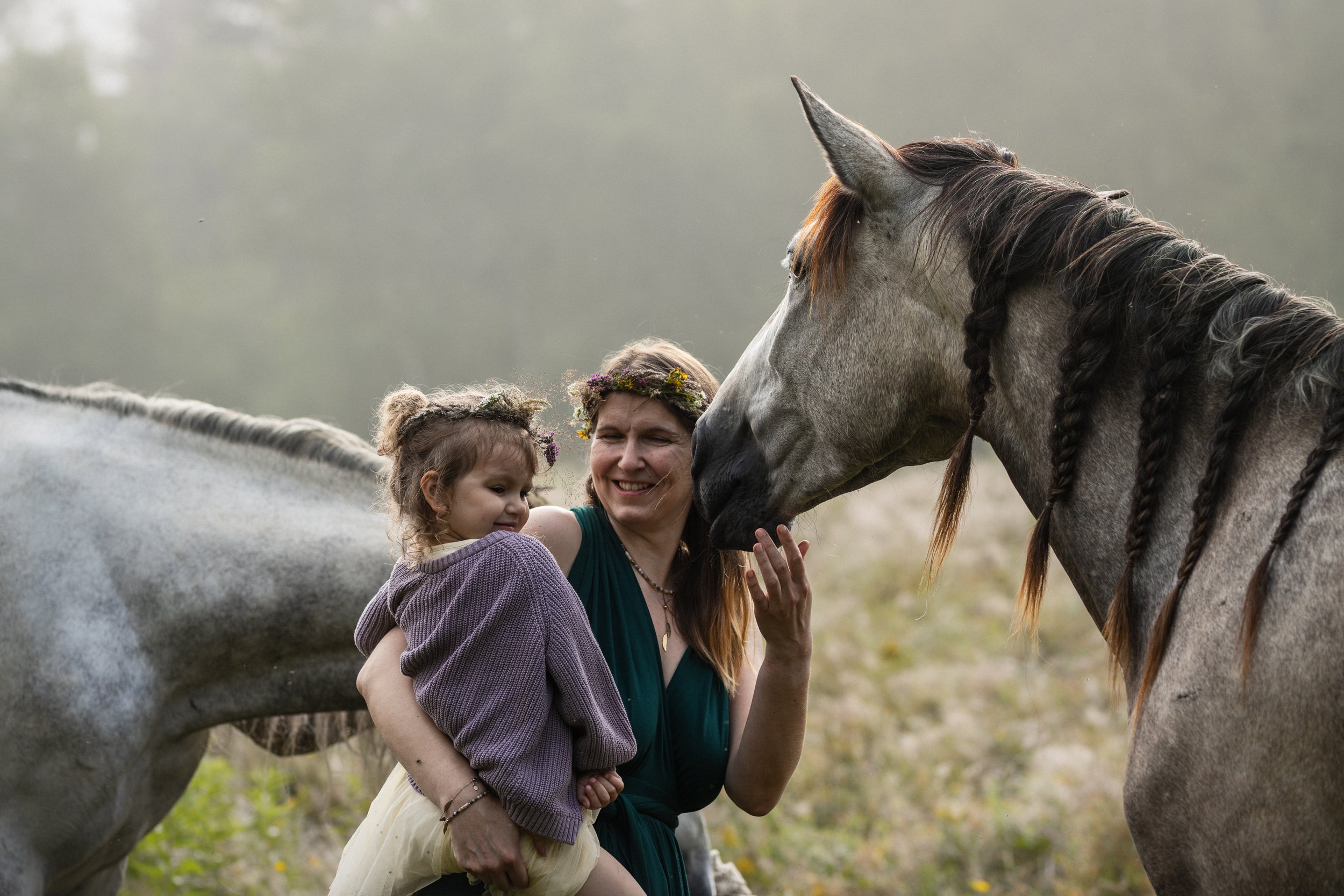 A woman with two children petting and hugging a gray horse outdoors in a natural setting.