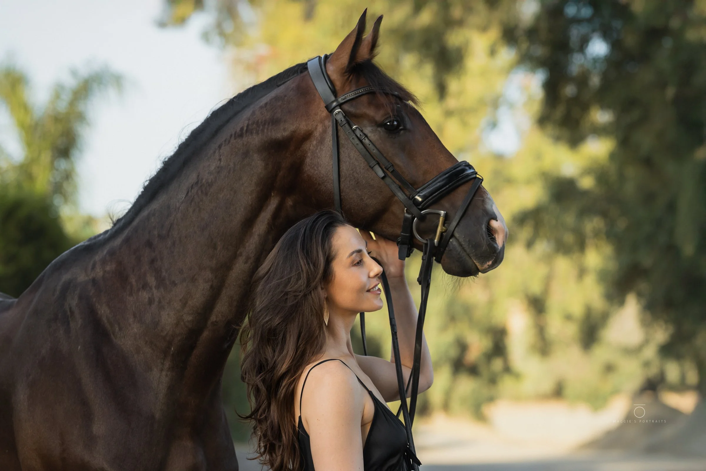 A woman with long brown hair and a black dress holding the bridle of a brown horse with a black mane outdoors with greenery in the background.