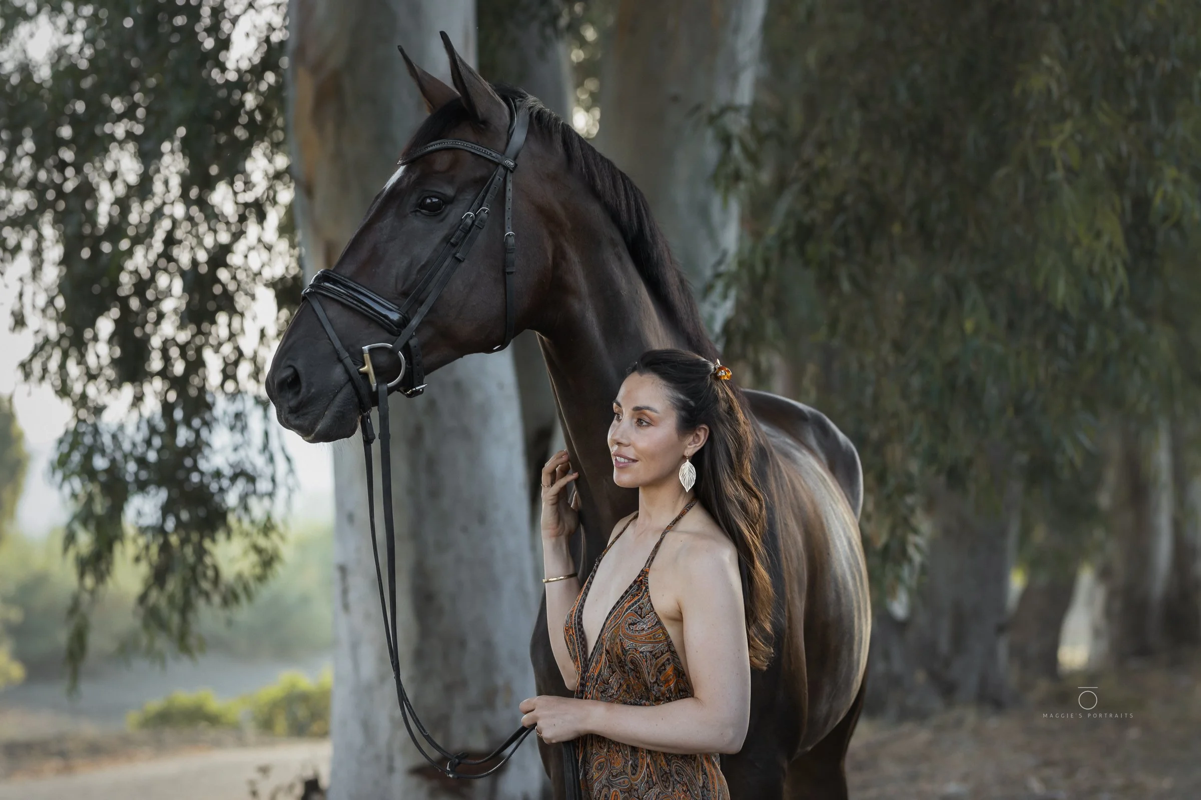 A woman with long dark hair wearing a patterned dress and earrings is standing next to a black horse with a bridle, touching its face in an outdoor setting with trees in the background.