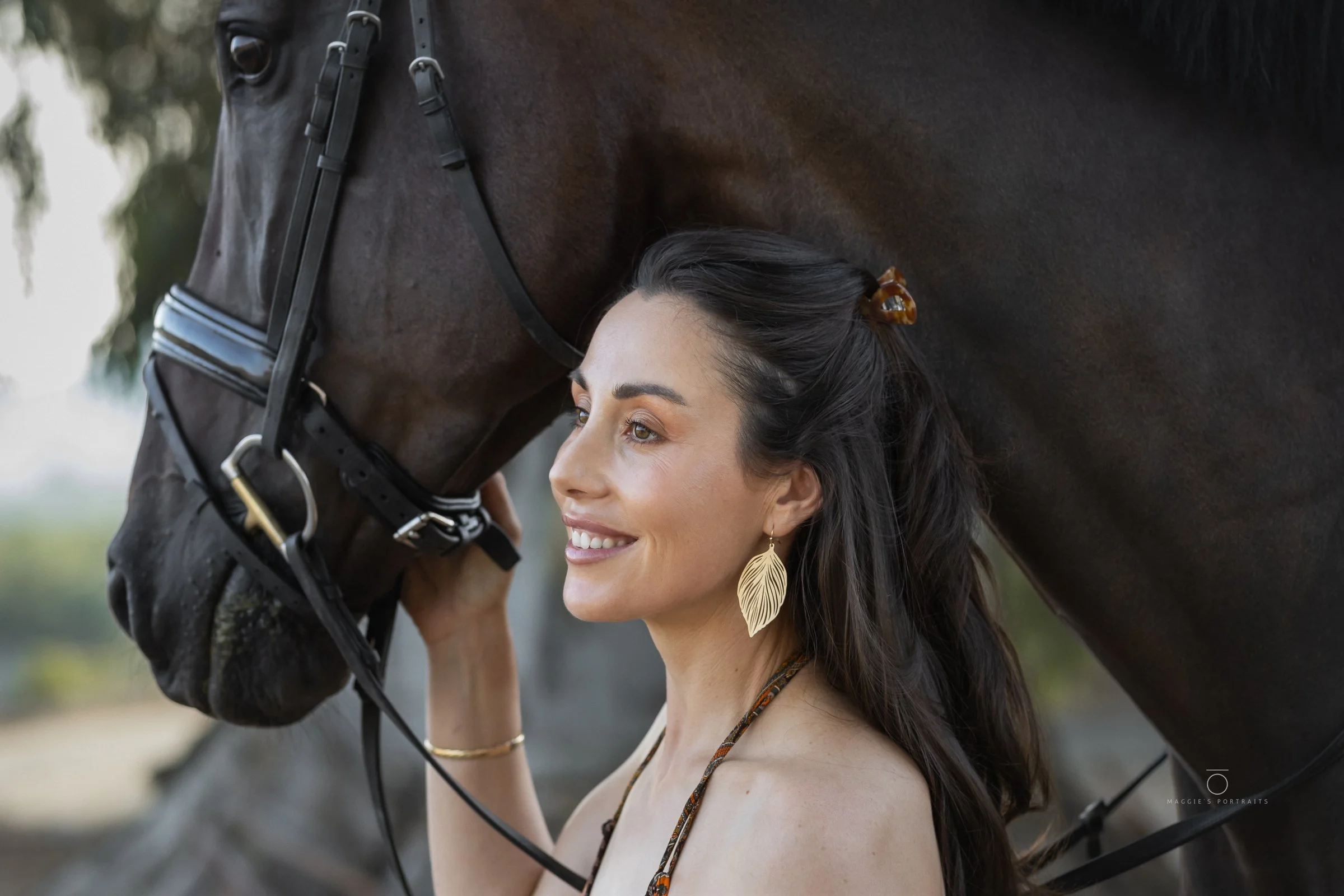 A woman with long dark hair and earrings smiling as a dark brown horse gently touches her forehead with its nose.