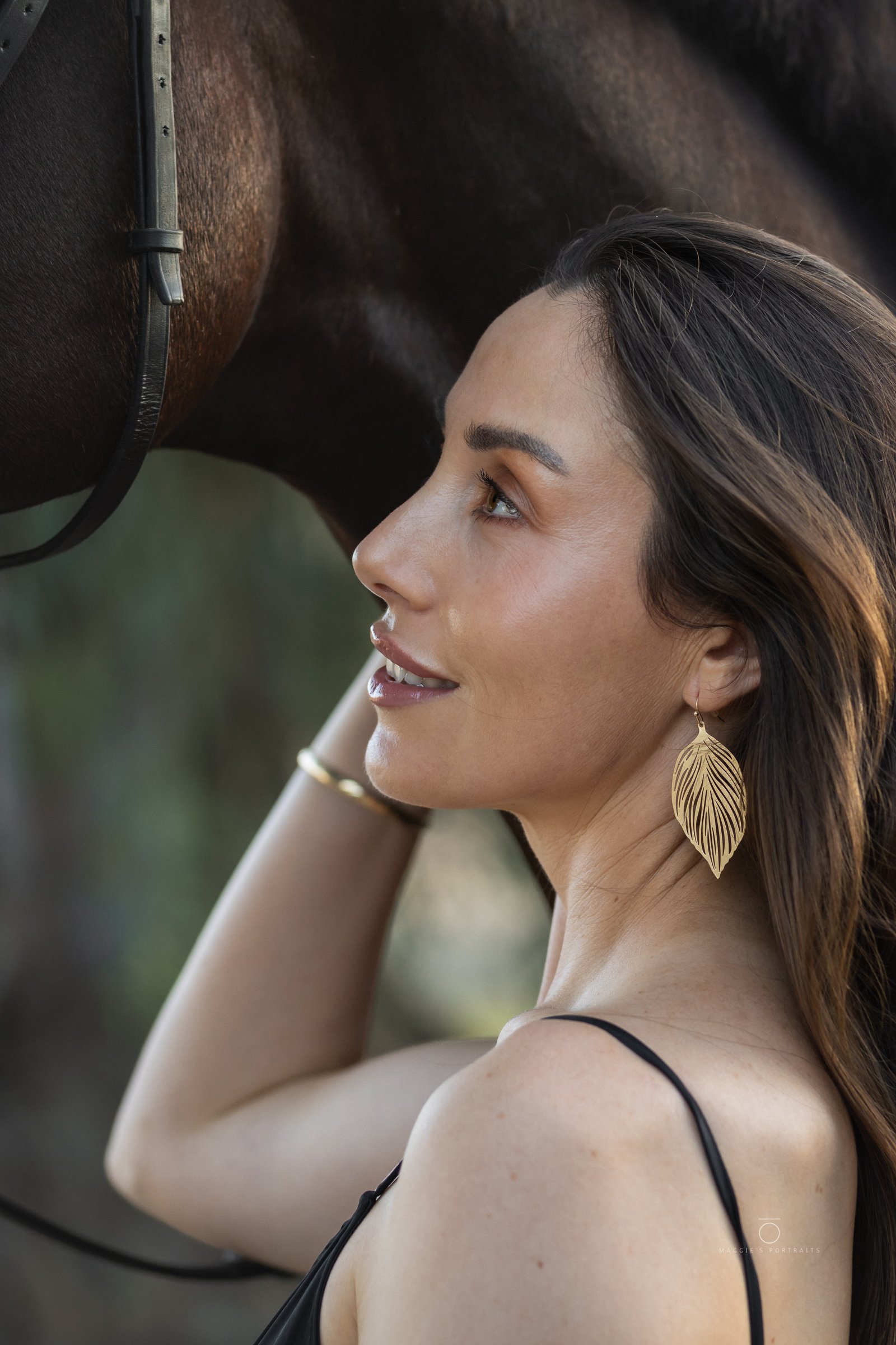 A woman with long brown hair and gold feather-shaped earrings, wearing a black spaghetti strap top, is resting her forehead against a dark brown horse wearing a bridle.