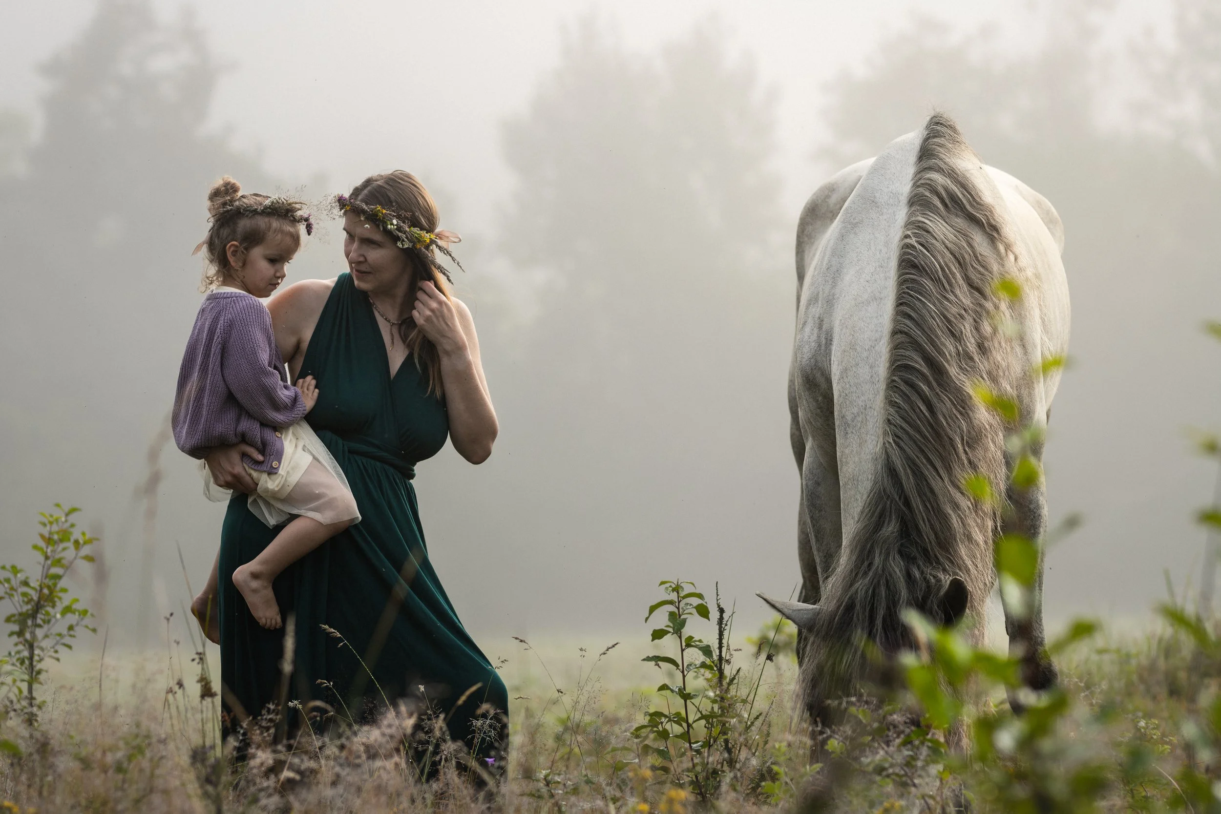 A woman wearing a dark green dress and a floral headband carries a young girl in a purple sweater and cream-colored skirt while walking through a foggy field next to a white horse.