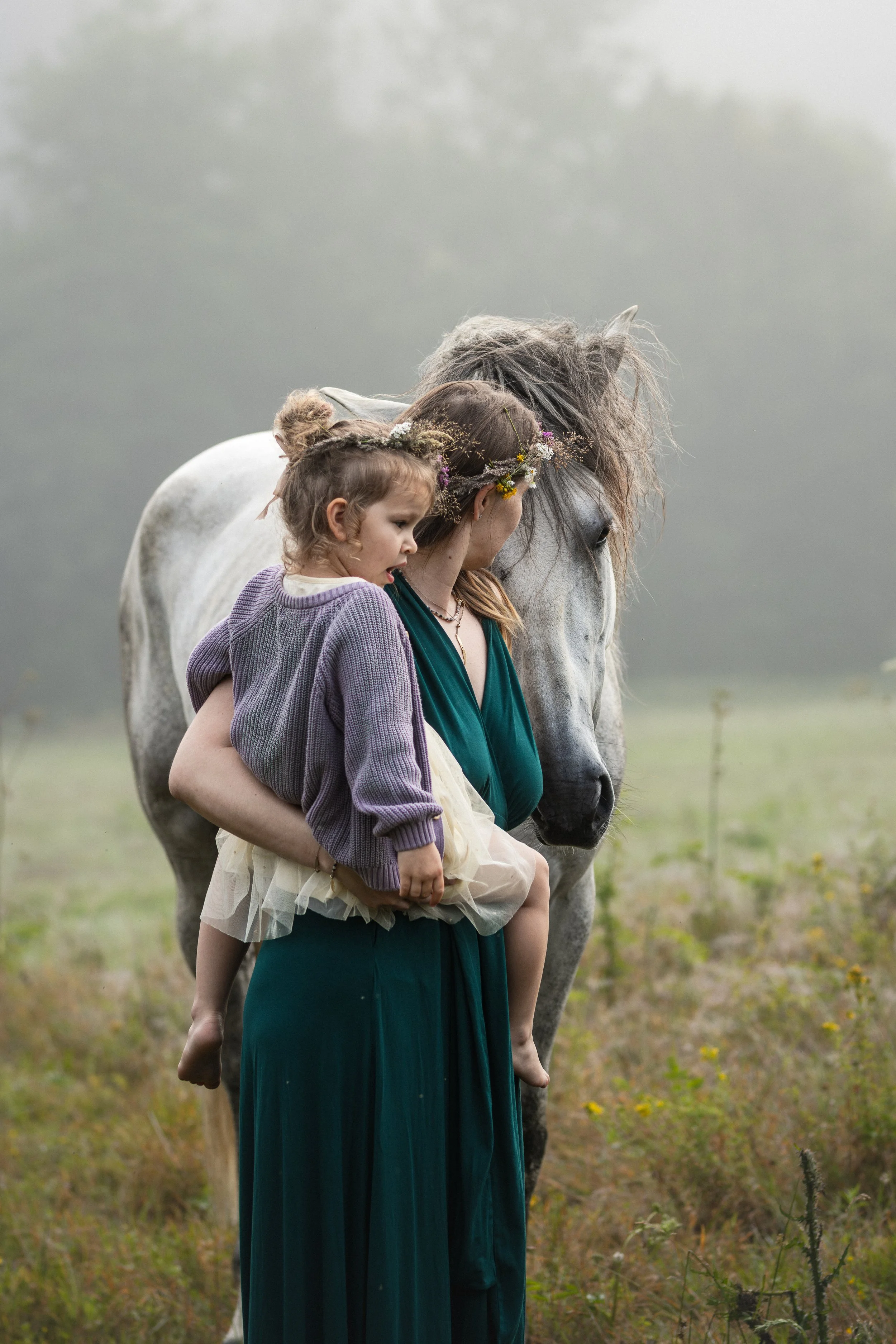 A woman with a flower crown and a girl with a bun hairstyle sit on a white horse with a gray mane. They are in a green field with yellow flowers, under an overcast sky.