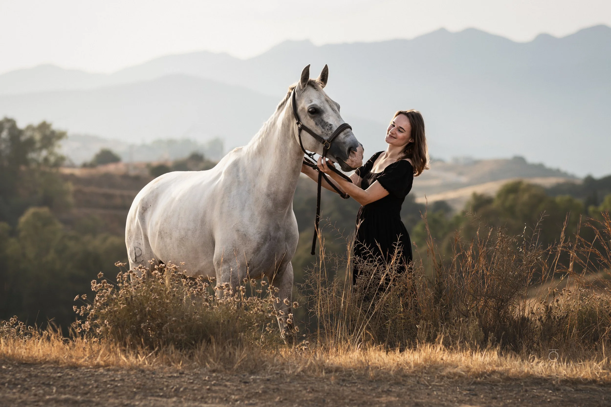 A woman smiling and holding the bridle of a white horse in a field with tall dry grass, mountains in the background, during daylight.