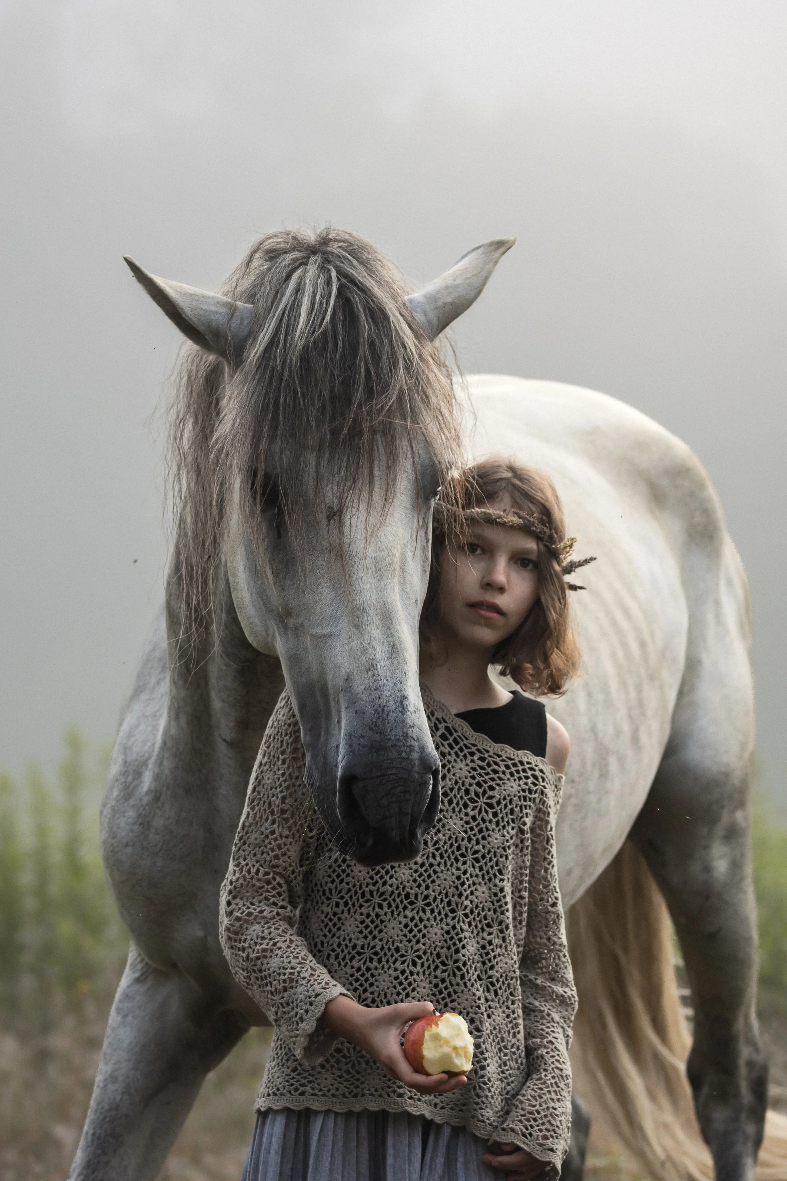 A young girl with curly brown hair, wearing a crochet sweater and a leafy headband, stands outdoors holding an apple with bitten portions. Next to her is a white horse with a gray mane, standing close with its head slightly lowered. The background is