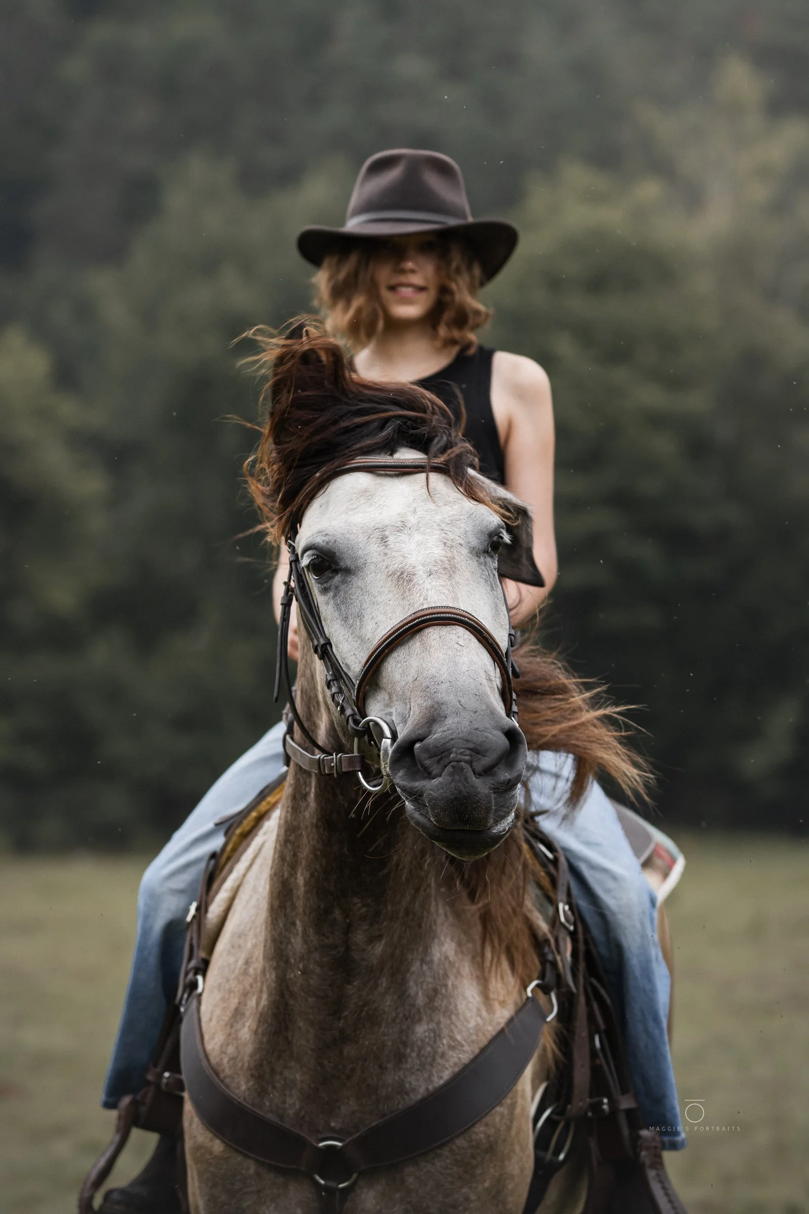 A woman wearing a wide-brimmed hat and black tank top riding a gray horse in a field with a blurred green background.