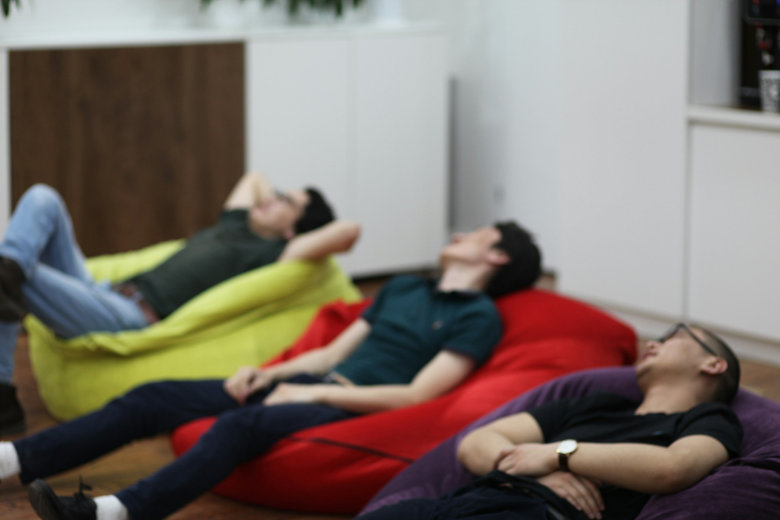 A group of young people is lying on their backs on colorful bean bags on the floor, relaxing with their eyes closed. They appear to be in a cozy indoor setting, possibly participating in a relaxing activity or meditation session.