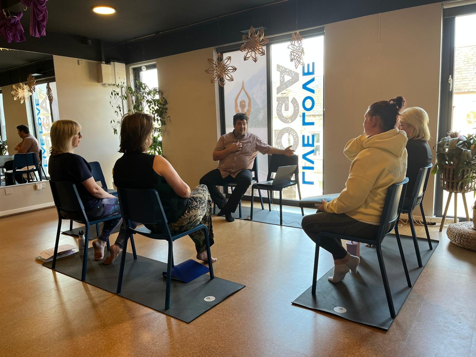 A group of five people seated in a semi-circle in a well-lit room, engaged in a discussion or workshop. One person appears to be leading the session, gesturing with their hand. The room has large windows, decorative hanging paper snowflakes, and a sign that reads ‘FIVE:CE’ on the window.