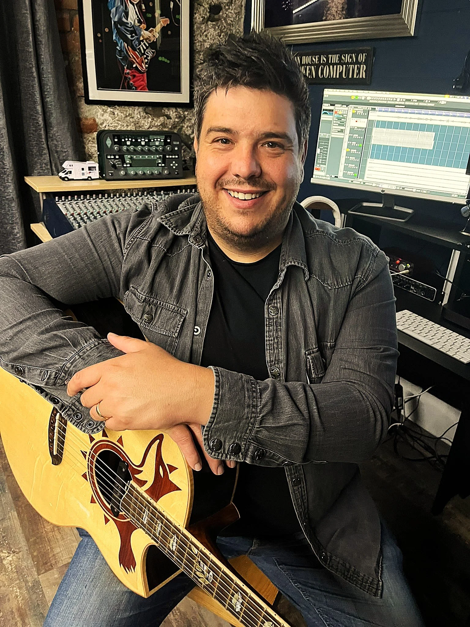 A smiling man with dark hair seated in a recording studio, holding a yellow acoustic guitar with a dragon design on the body. Behind him are music production equipment, a computer monitor displaying music software, and framed pictures on the wall.