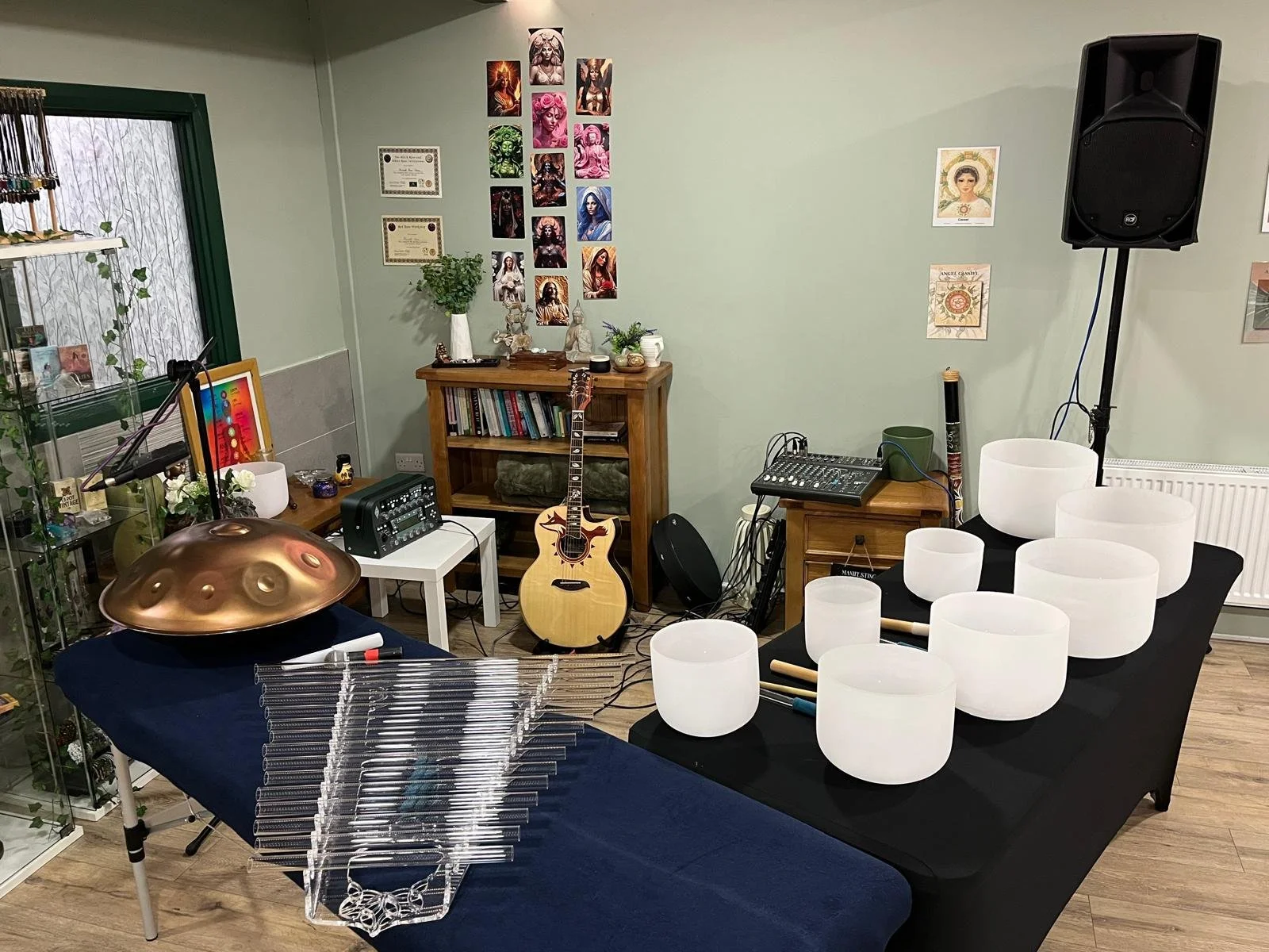 A room set up for a musical performance with various instruments including a hang drum, crystal singing bowls, a guitar, a chime, and audio equipment. The walls are decorated with artwork and framed certificates, and there is a window with green trim and a display of jewelry.