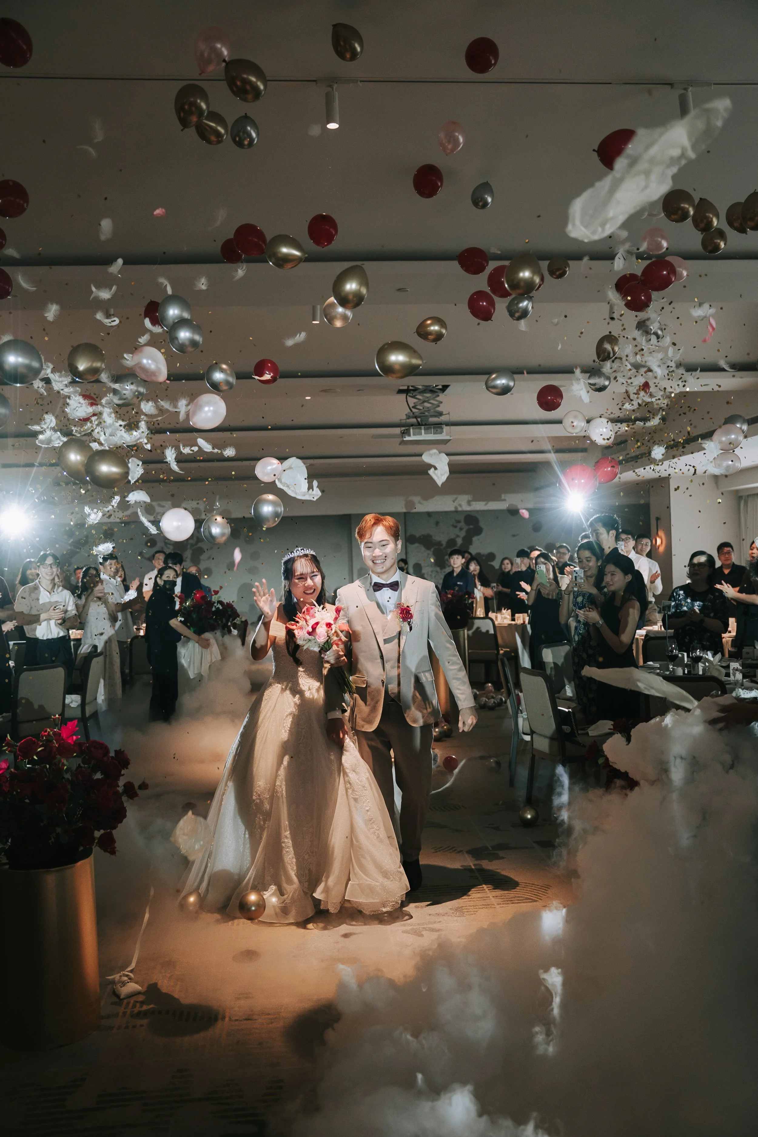 A bride and groom smiling and holding hands as they walk into a celebration, surrounded by guests, balloons, and confetti, with fog and dramatic lighting.